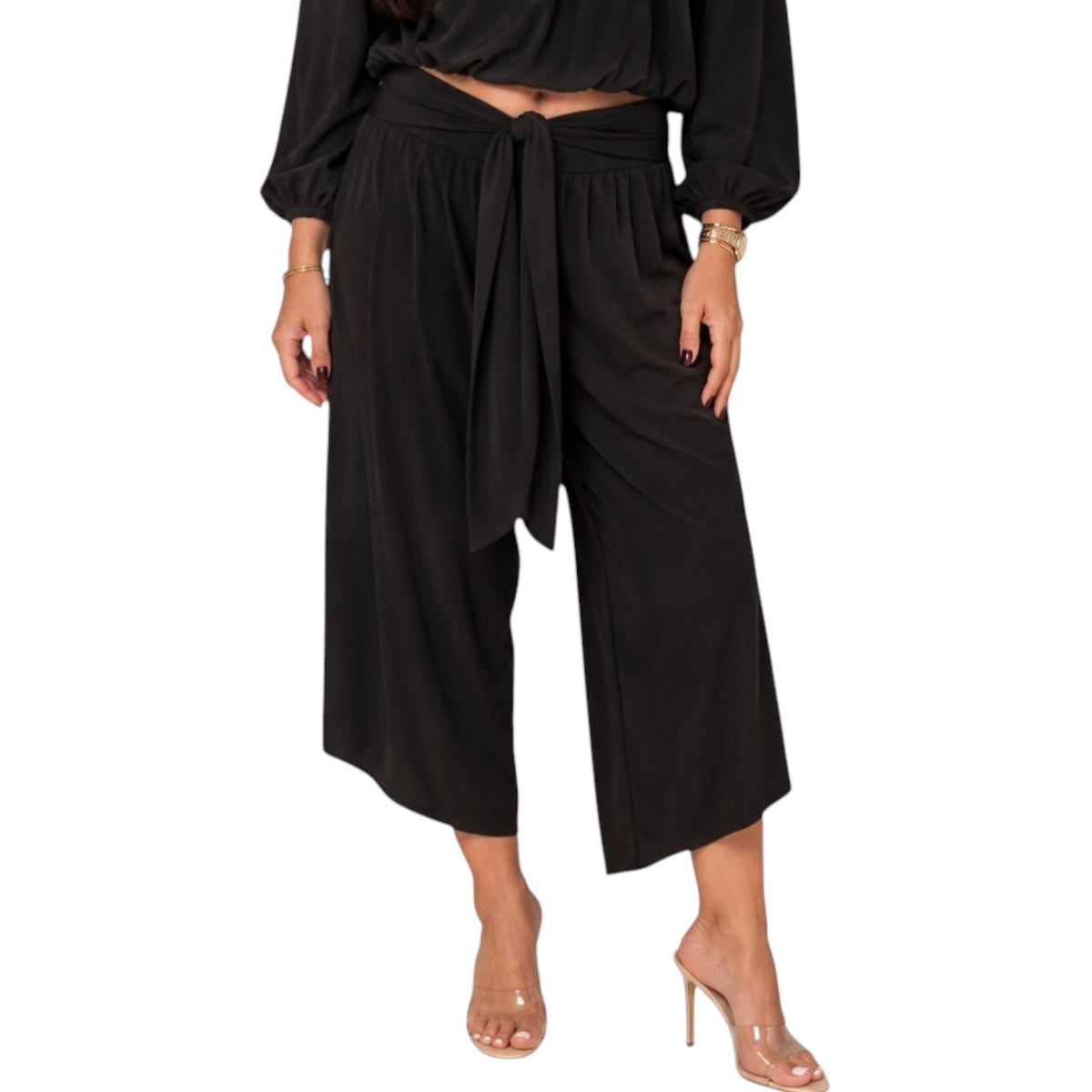 A model wearing black wide-leg cropped pants with a tie waist, paired with a black long-sleeve crop top and clear high-heeled sandals, shown against a plain white background.