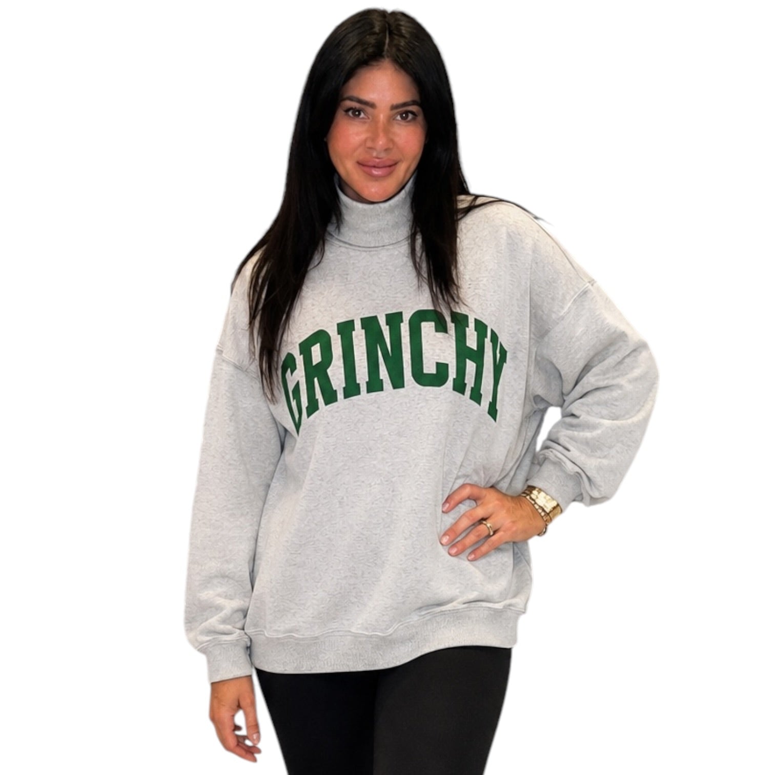 A woman modeling a light gray turtleneck sweatshirt with 'GRINCHY' printed in green arched letters across the chest, paired with black leggings, against a plain white background.