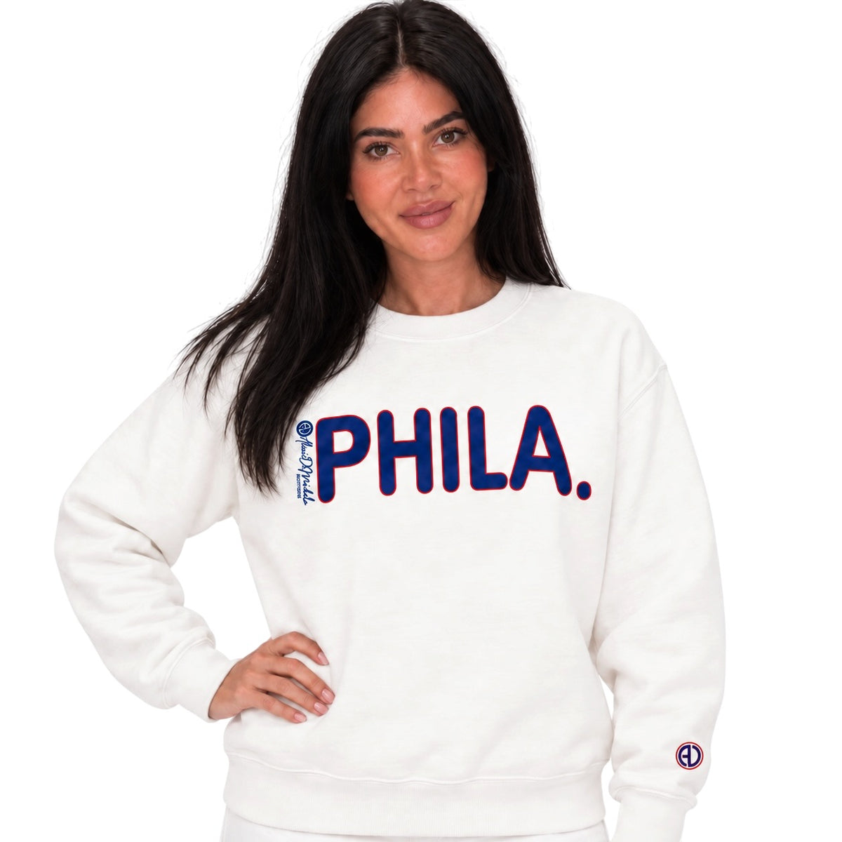 A woman modeling a white crewneck sweatshirt with 'PHILA.' printed across the chest in navy blue with a red outline, and a small circular logo on the left sleeve.