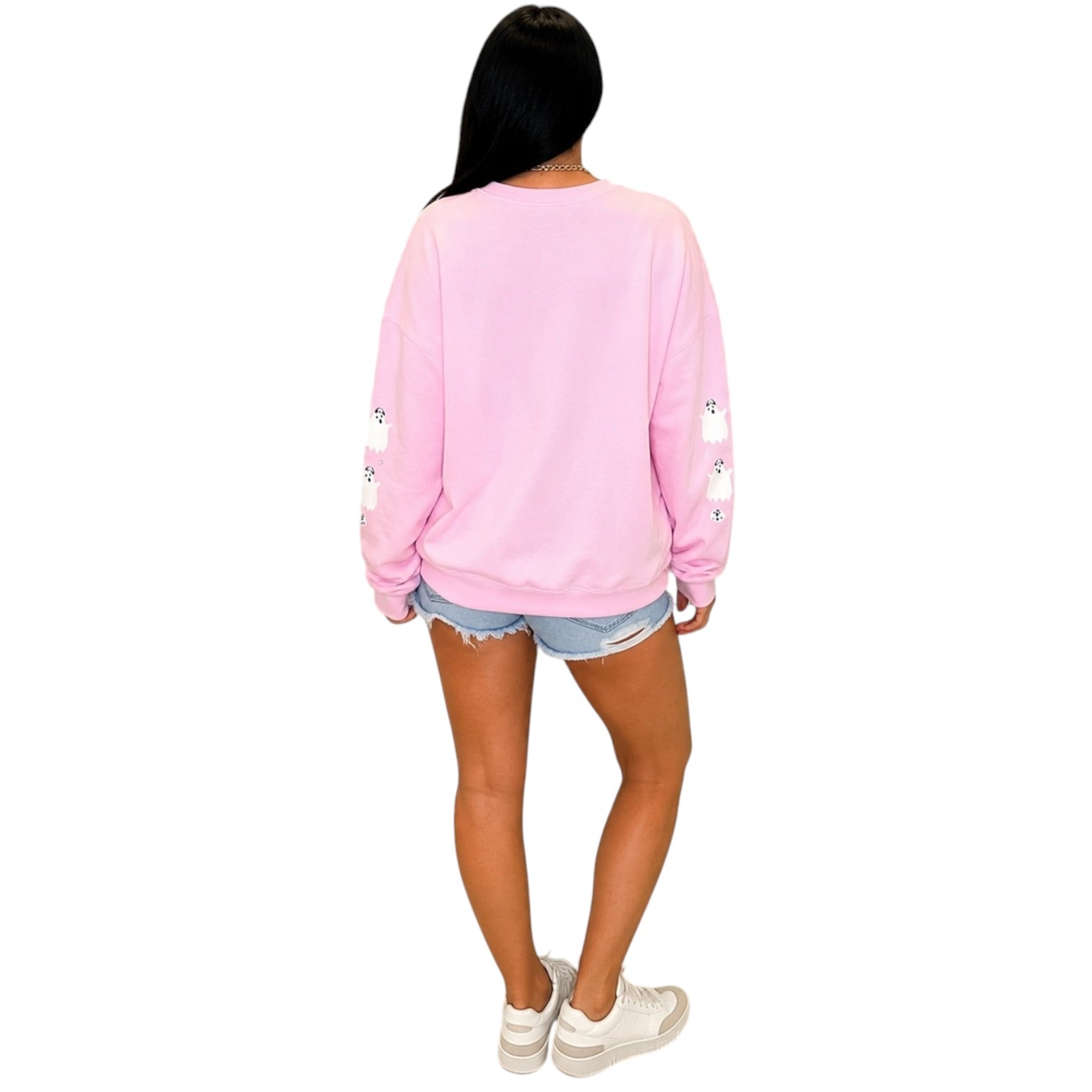 A woman shown from the back wearing a light pink crewneck sweatshirt with small white cartoon graphics on the sleeves, paired with denim shorts and white sneakers, against a plain white background.