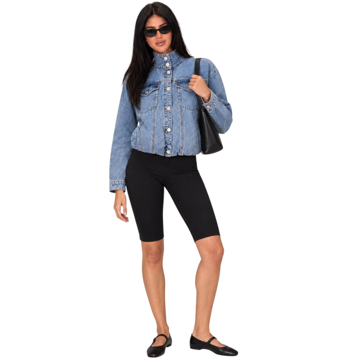 A woman modeling a light-wash blue denim jacket, black biker shorts, black flats, and carrying a black shoulder bag against a plain white background.
