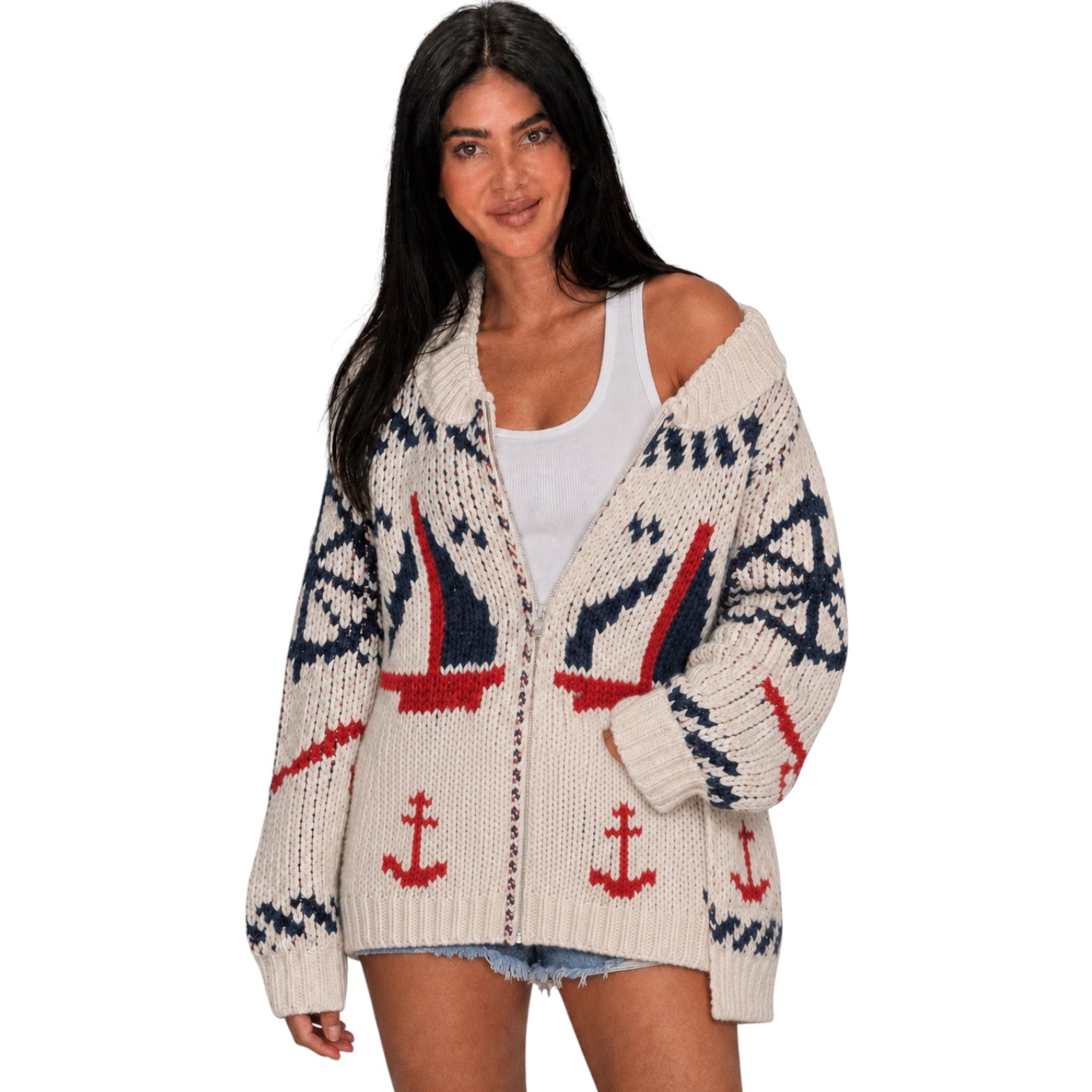 A woman modeling a cream knit cardigan with a nautical pattern featuring red and navy blue sailboats, anchors, and compass designs, worn over a white tank top.