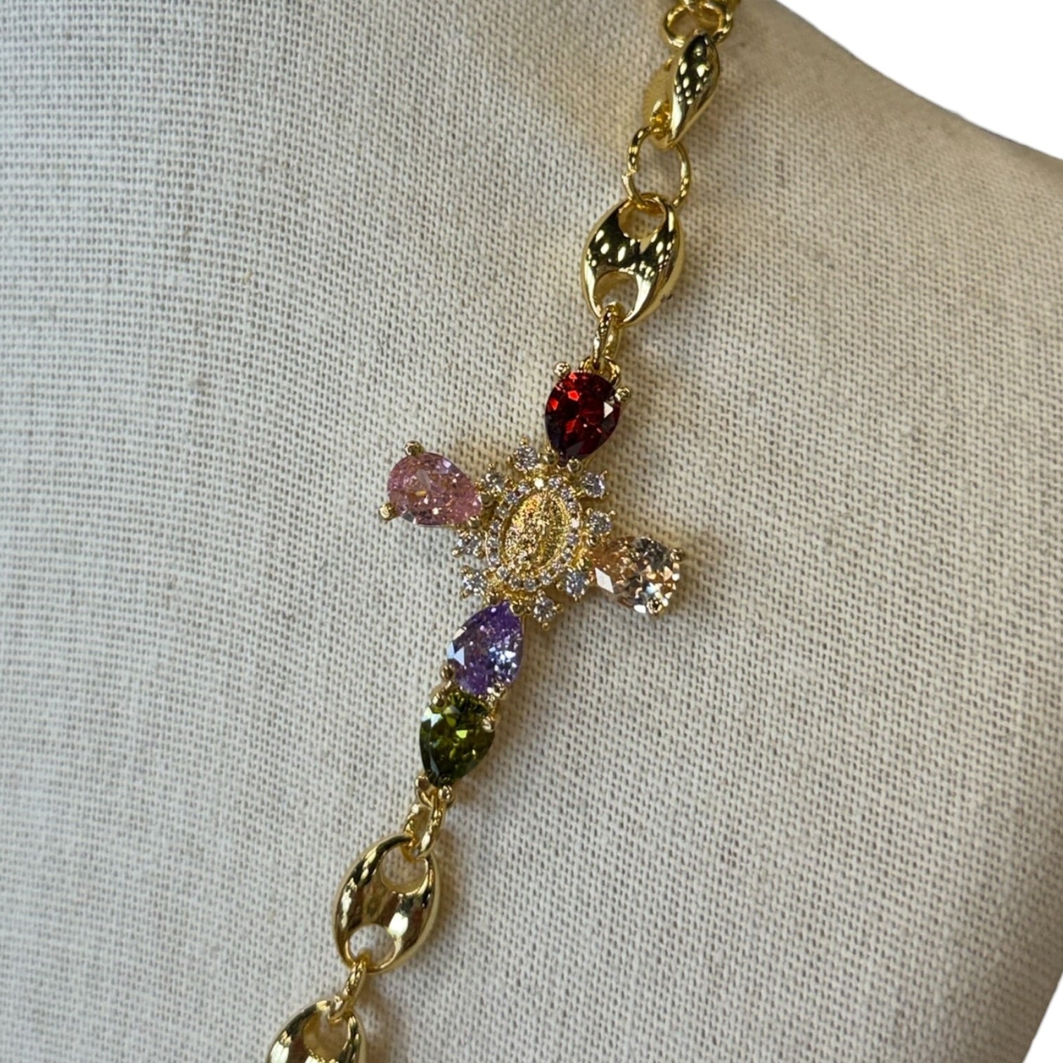 A gold-tone cross necklace with multicolored gemstone accents, displayed on a beige fabric jewelry bust.