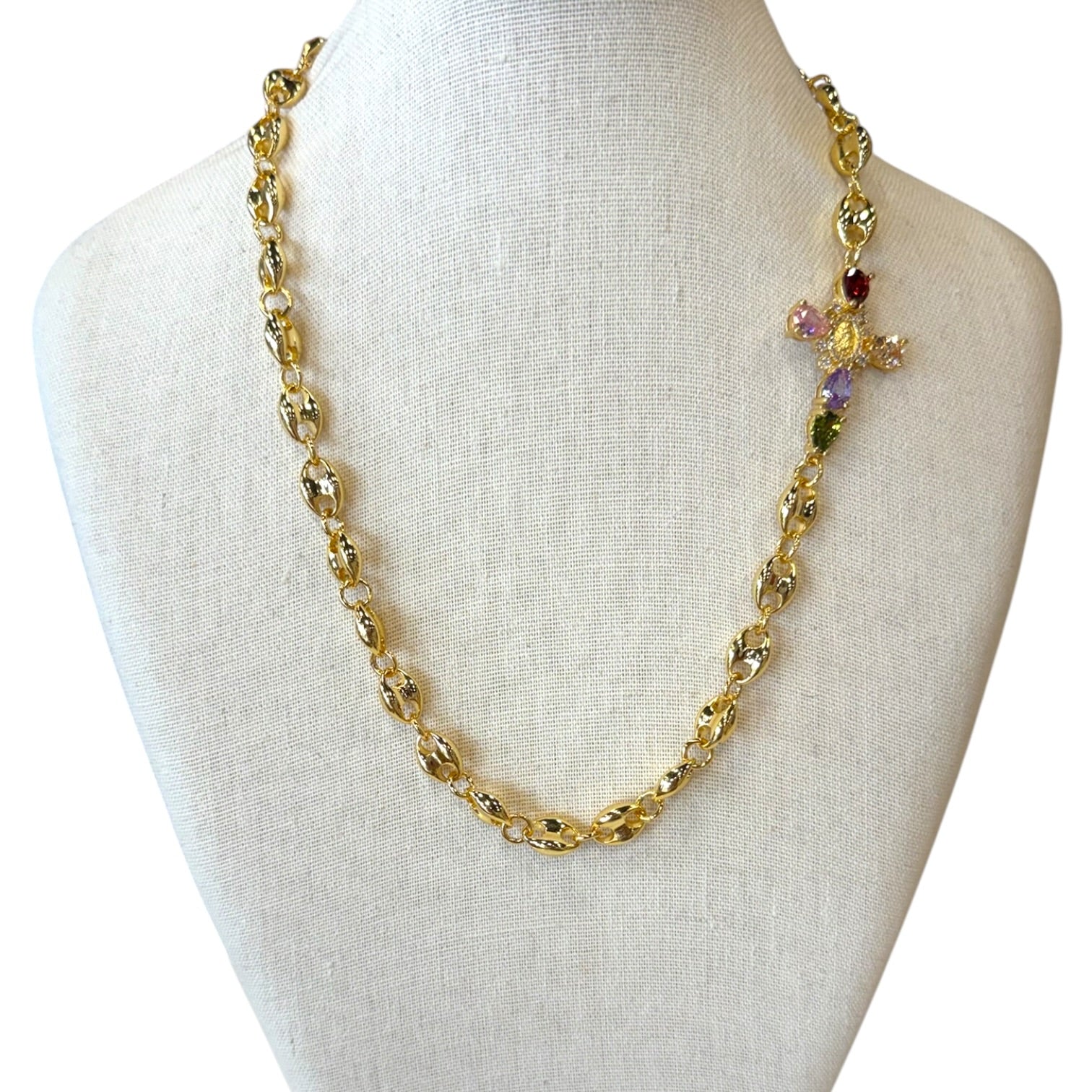 A gold-toned chain necklace displayed on a cream fabric jewelry bust. The necklace features textured oval links and a small decorative cross charm embellished with pastel purple, green, and red stones.