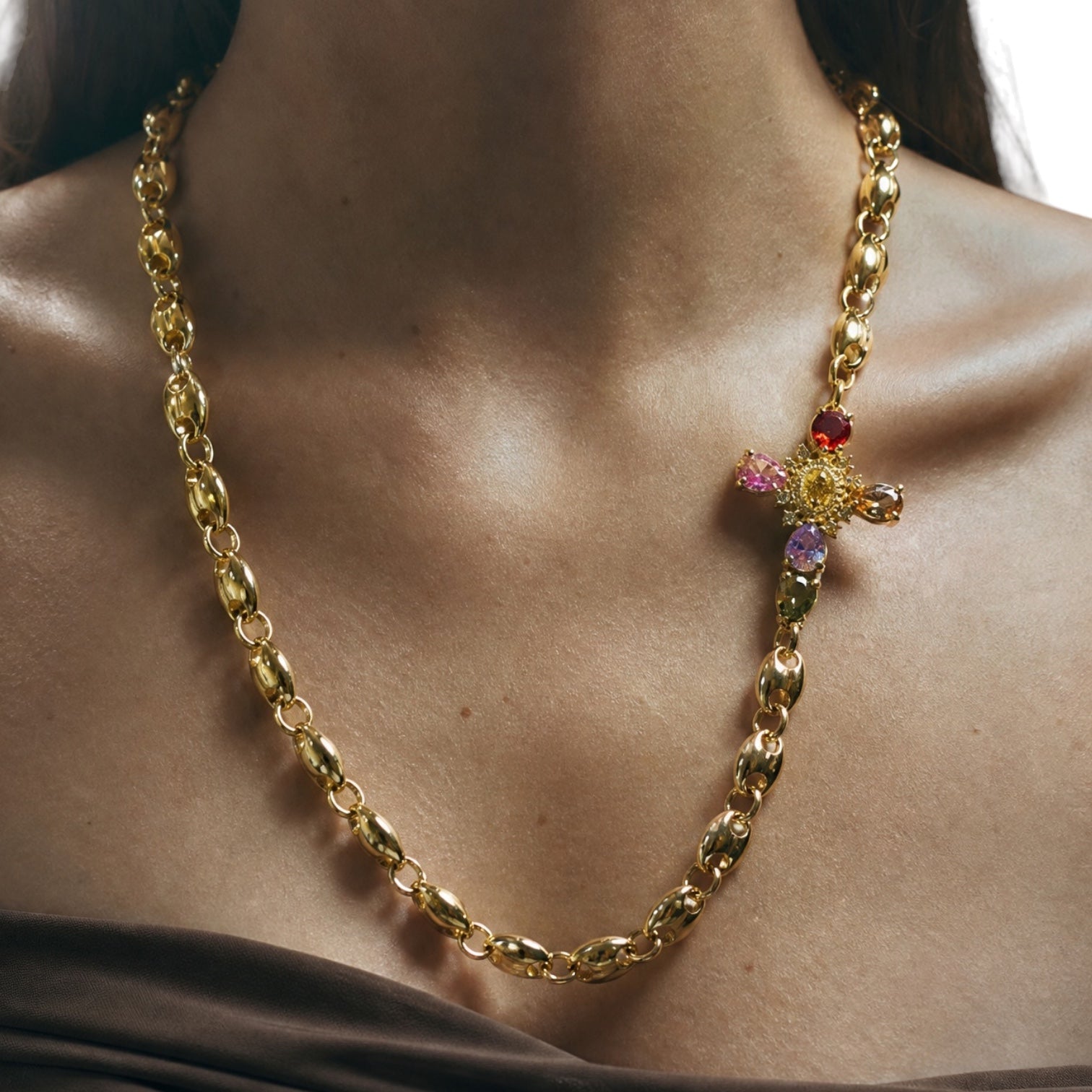 A gold chain necklace with a decorative cross pendant featuring pink, purple, and red gemstones, worn on a model's neck.