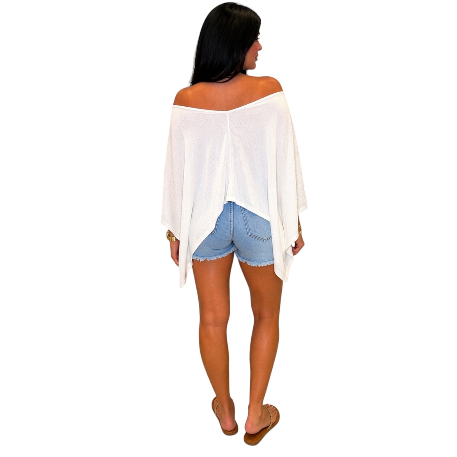 A woman shown from the back wearing a white off-the-shoulder poncho top, light blue denim shorts, and brown flat sandals against a plain white background.