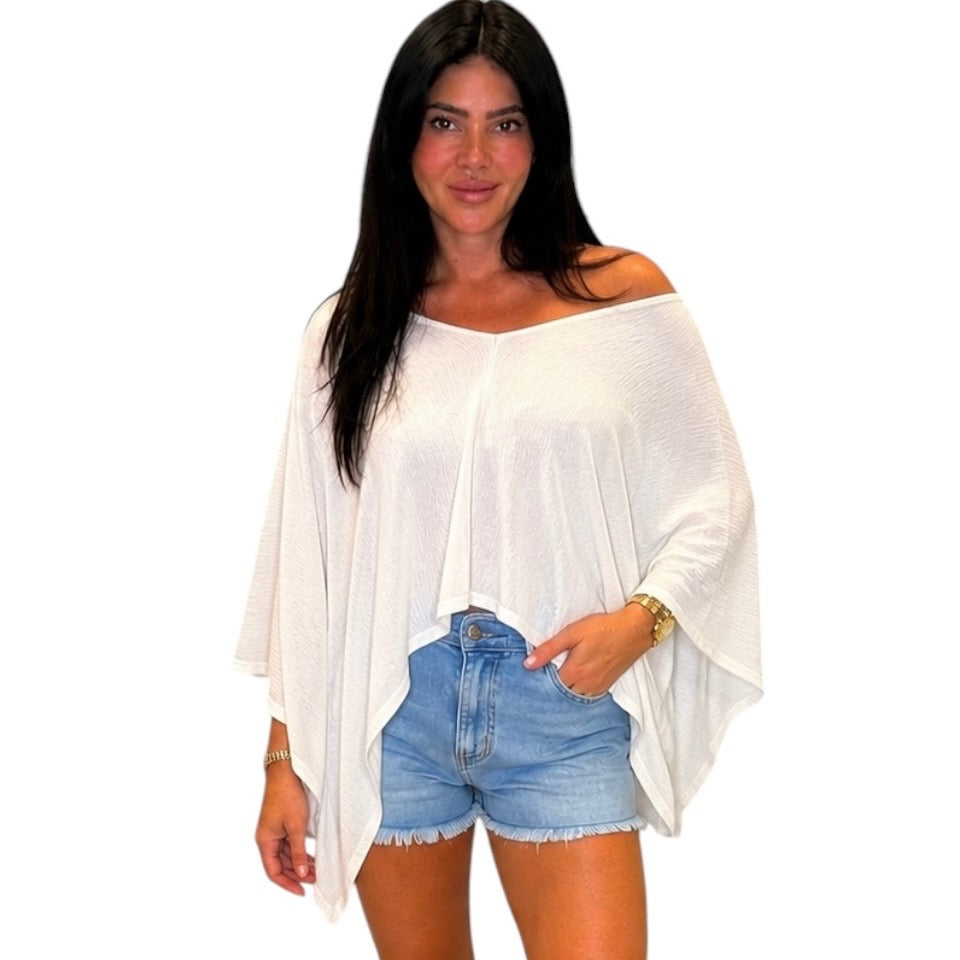 A woman modeling a white off-the-shoulder, long-sleeve poncho-style top paired with light blue denim shorts against a plain white background.