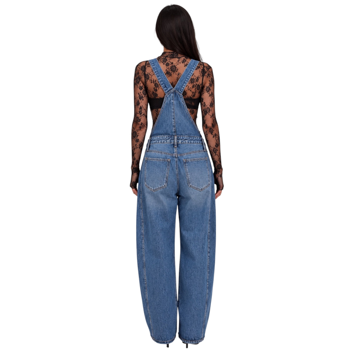 A woman viewed from the back wearing blue denim overall-style pants with wide legs and a black sheer floral lace long-sleeve top, against a plain white background.