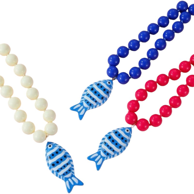 Three beaded necklaces on a white background: one with cream beads, one with bright blue beads, and one with bright red beads. Each necklace features a blue and white striped fish-shaped pendant.
