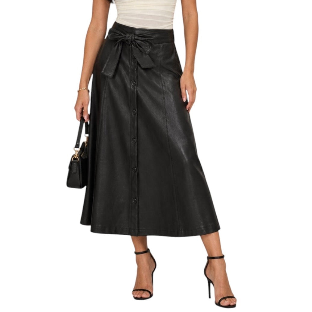A model wearing a black faux-leather midi skirt with a tie waist and front button closure, paired with a white ribbed top and black high-heeled sandals, against a plain white background.