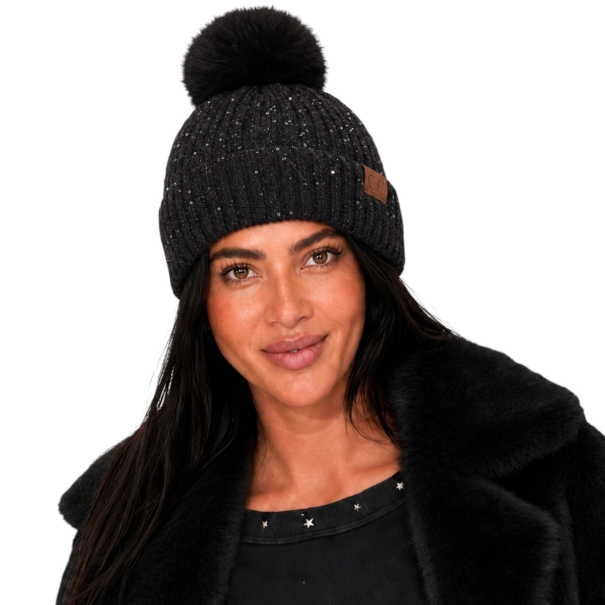 A woman wearing a black knit beanie with a large black pom-pom and subtle silver speckles, paired with a black faux-fur collar jacket. The beanie features a small brown leather brand patch on the side.