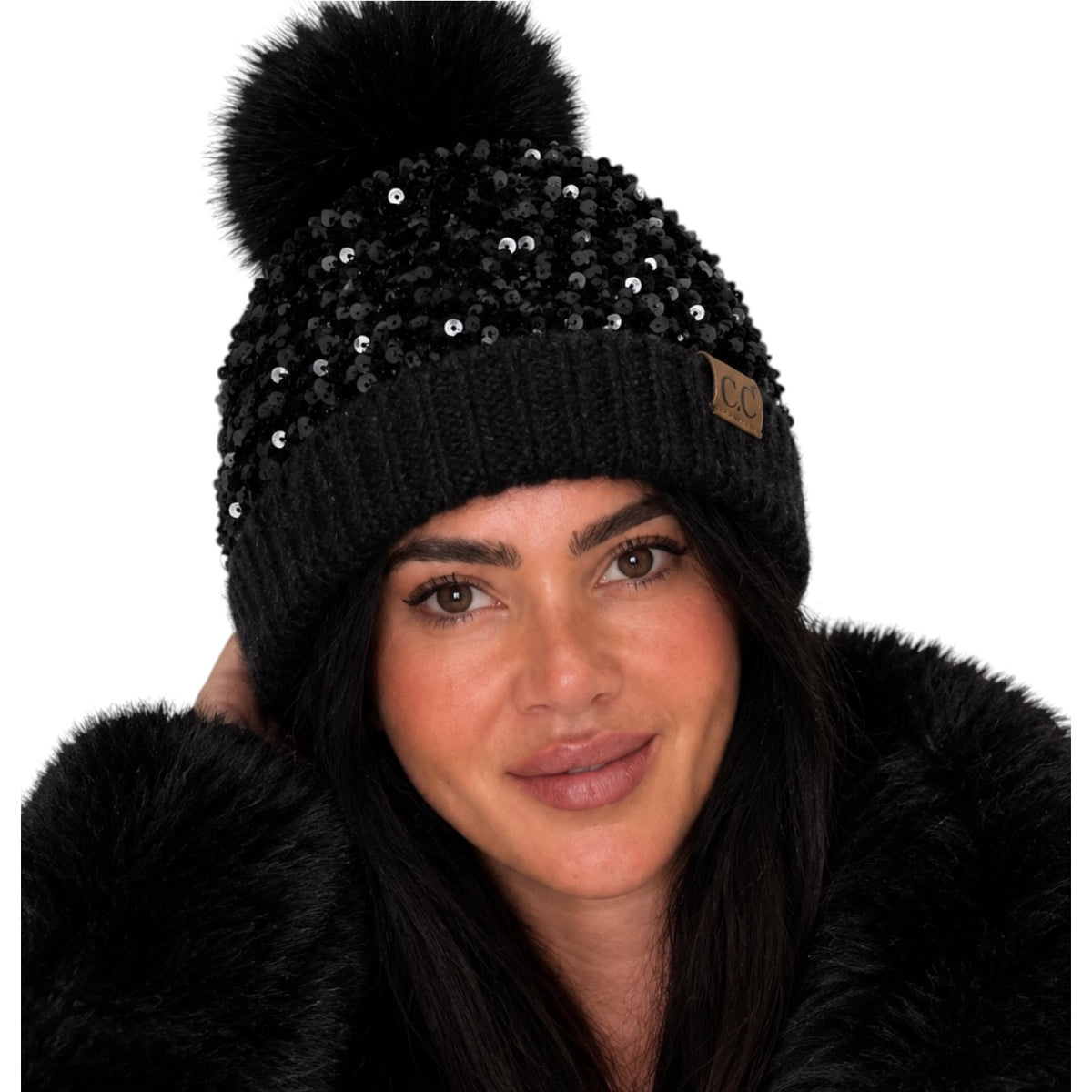 A close-up of a woman wearing a black knit beanie with a large black faux-fur pom-pom, black sequins, and a small brown UGG logo patch on the side.
