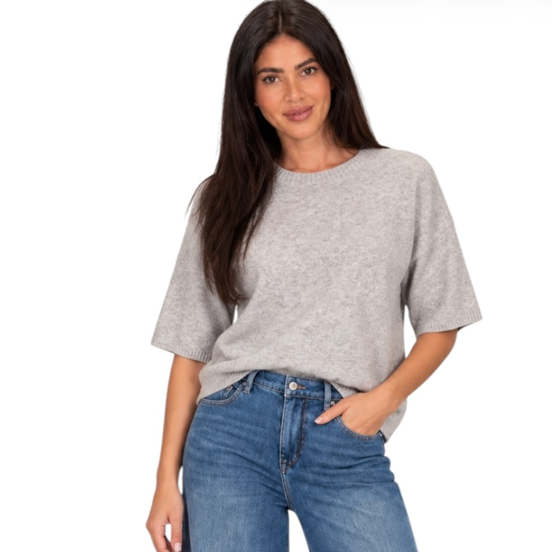 A woman modeling a light gray short-sleeve knit top with blue jeans against a plain white background.