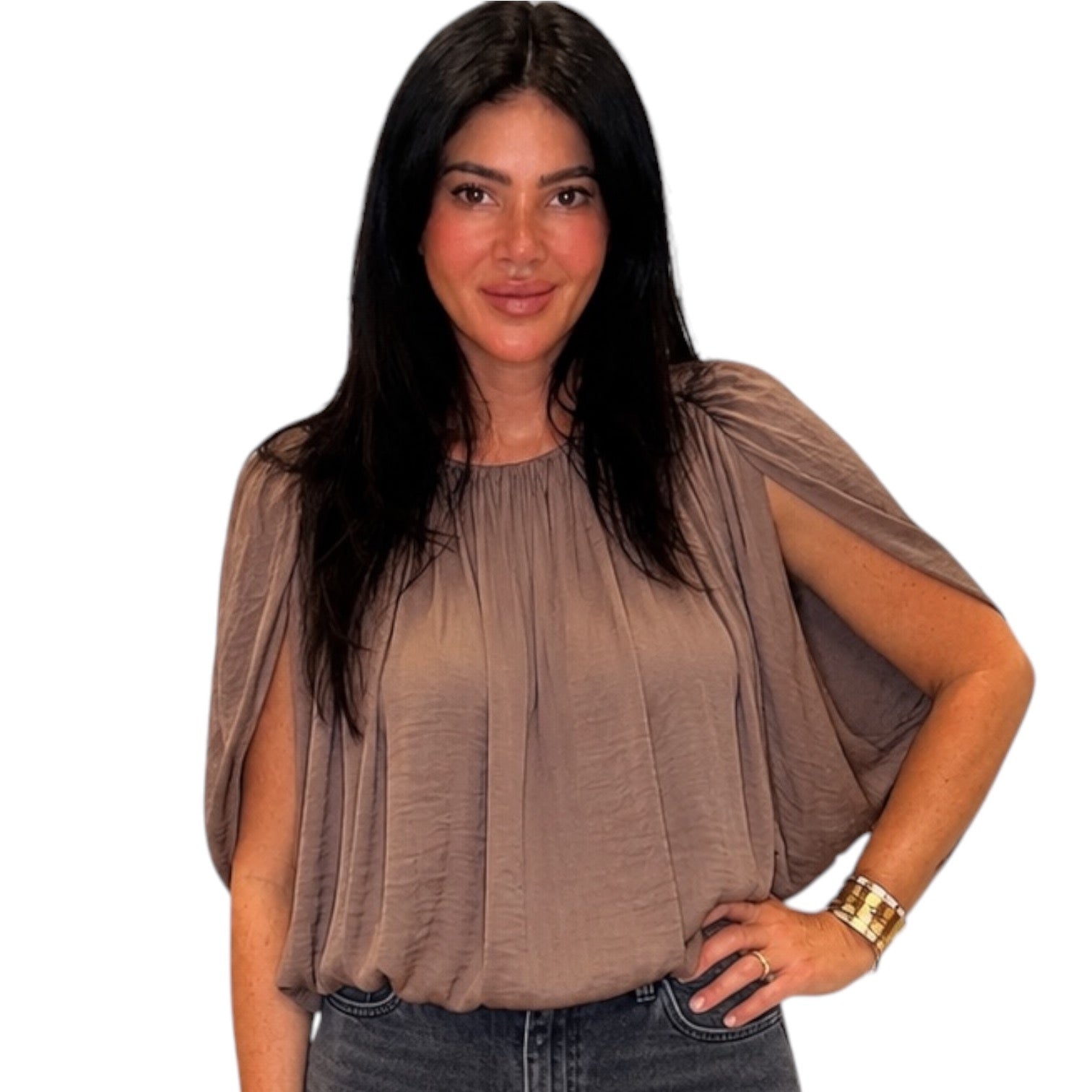 A woman with long black hair wearing a taupe-brown draped short-sleeve blouse and dark jeans, standing against a plain white background.