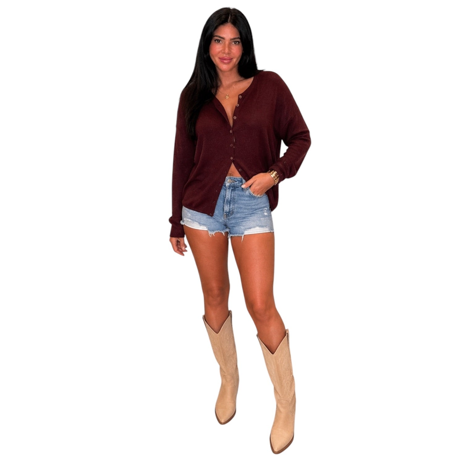 A woman modeling a long-sleeve burgundy button-up top, paired with light-wash denim shorts and beige cowboy boots, against a plain white background.