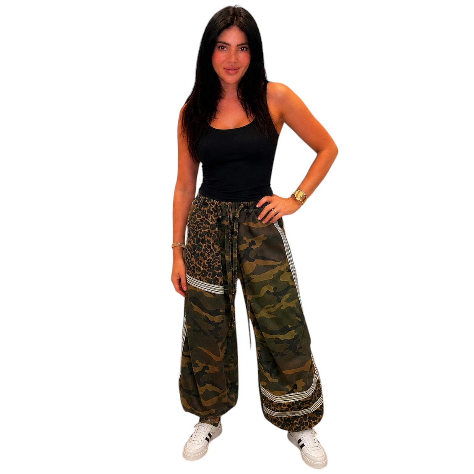 A woman modeling wide-leg harem pants with a green and brown camouflage pattern and leopard print side panels, paired with a black tank top and white sneakers, against a plain white background.