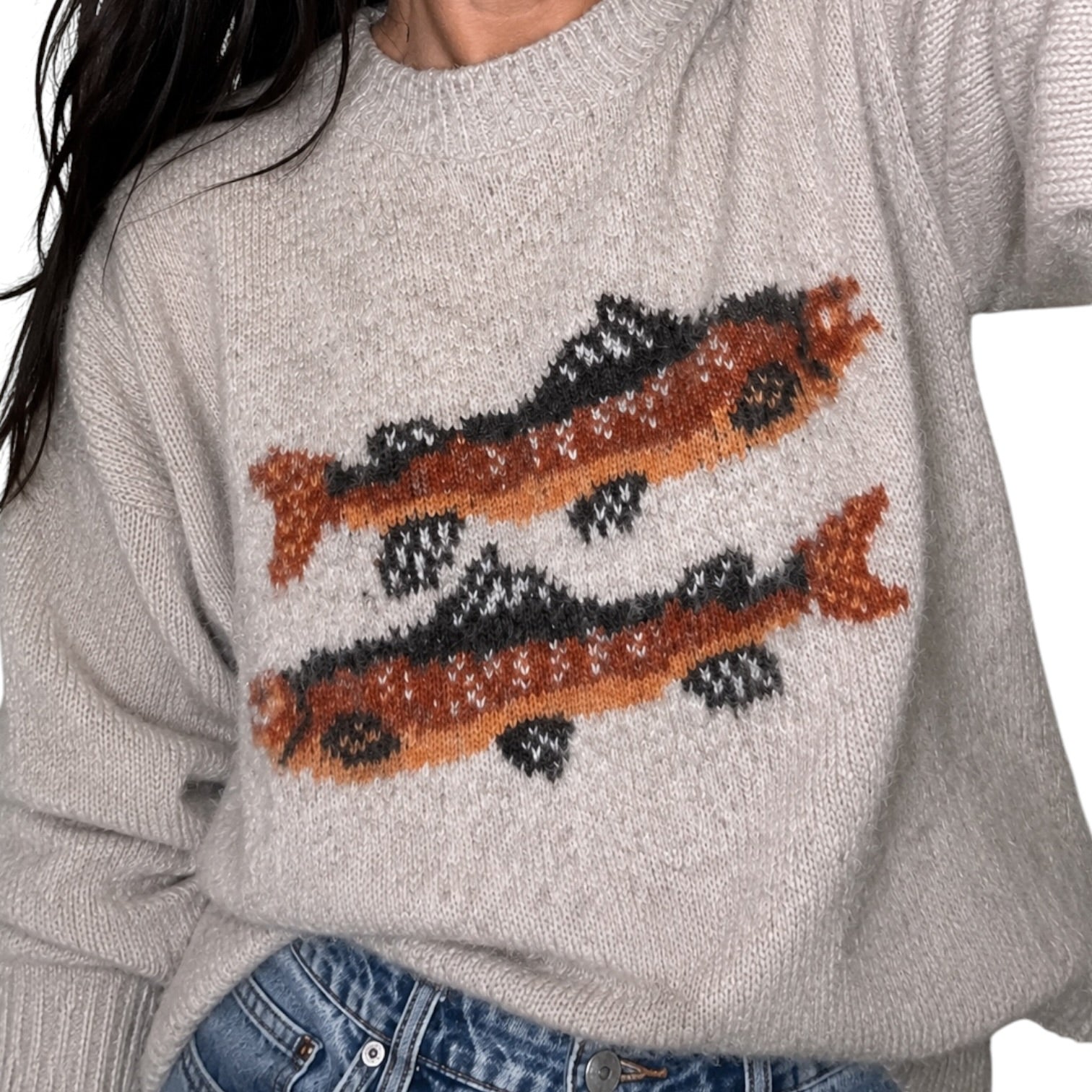 A close-up of a person wearing a light heather gray knit sweater with two large fish motifs in orange, black, and white across the chest, paired with blue jeans.