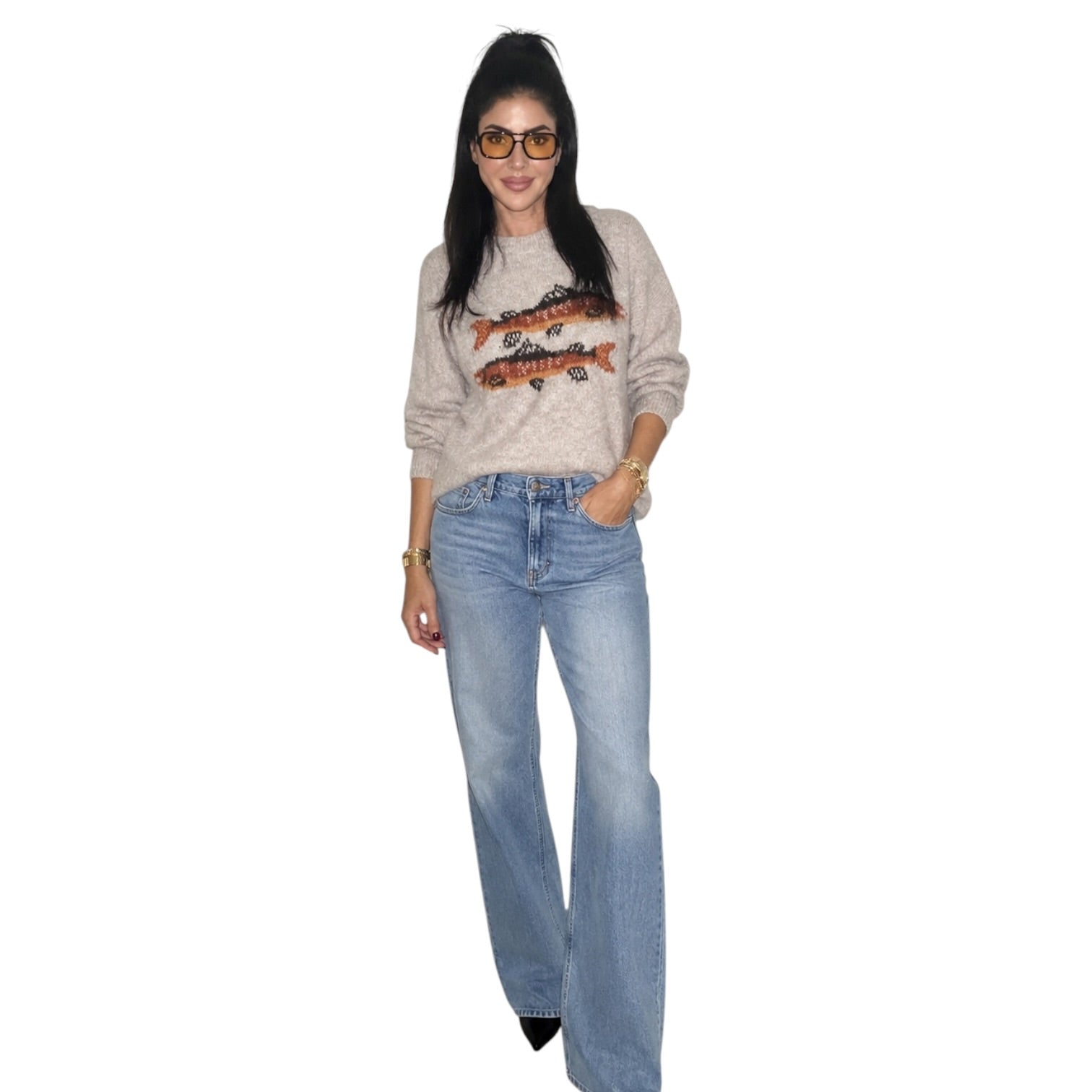 A woman modeling a beige crewneck sweatshirt with a fish graphic and light-wash blue jeans, standing against a plain white background.
