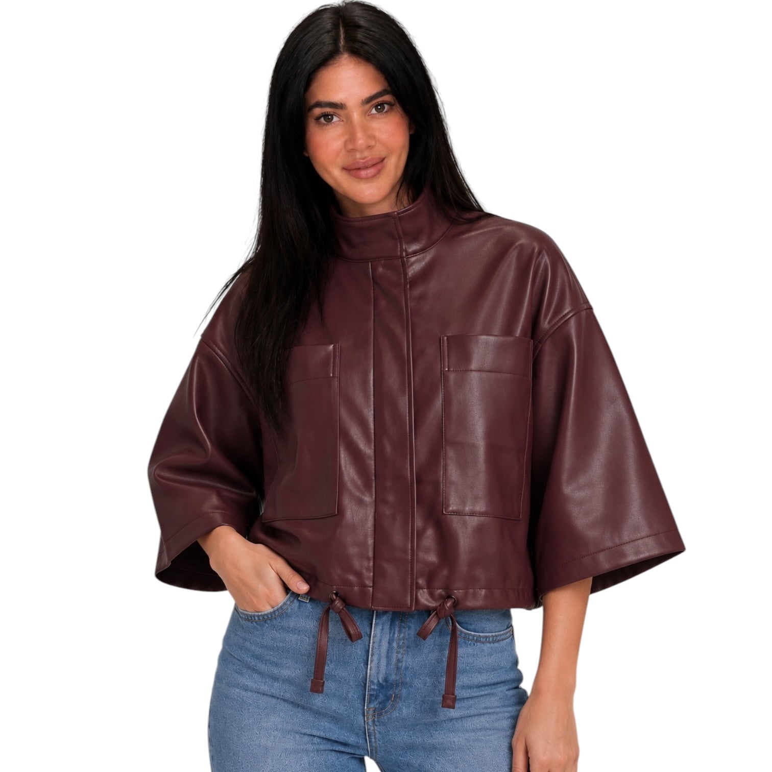 A woman modeling a cropped, dark burgundy faux-leather jacket with wide three-quarter sleeves, a high mock neck, and two large front patch pockets, paired with blue jeans against a plain white background.