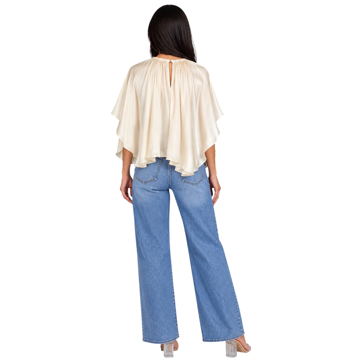 A woman shown from the back wearing a cream-colored, semi-sheer draped top with wide flutter sleeves and a keyhole back, paired with blue wide-leg jeans and clear high-heeled sandals, against a plain white background.