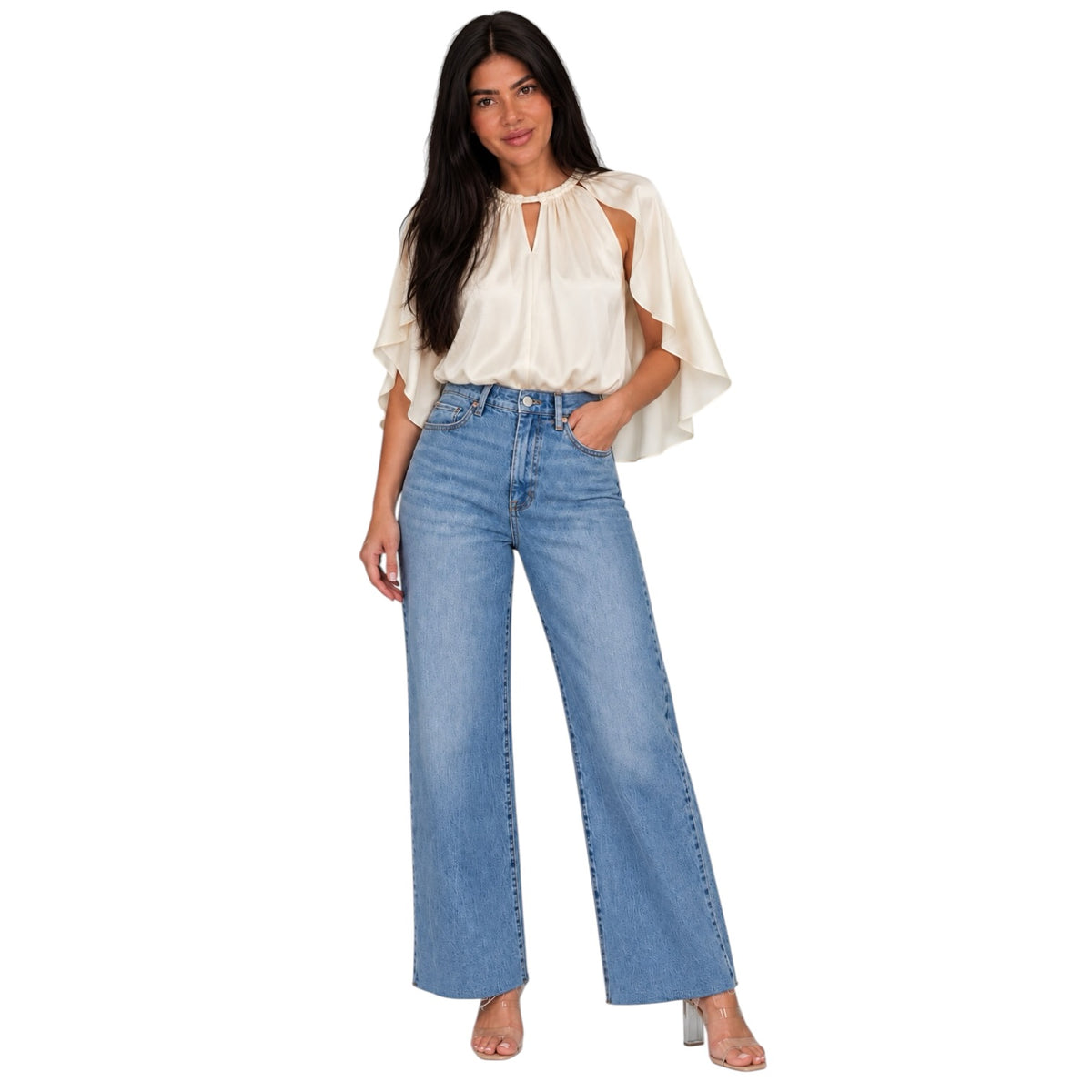 A woman modeling a cream-colored cold-shoulder blouse with ruffled sleeves and light-wash blue wide-leg jeans, standing against a solid white background.