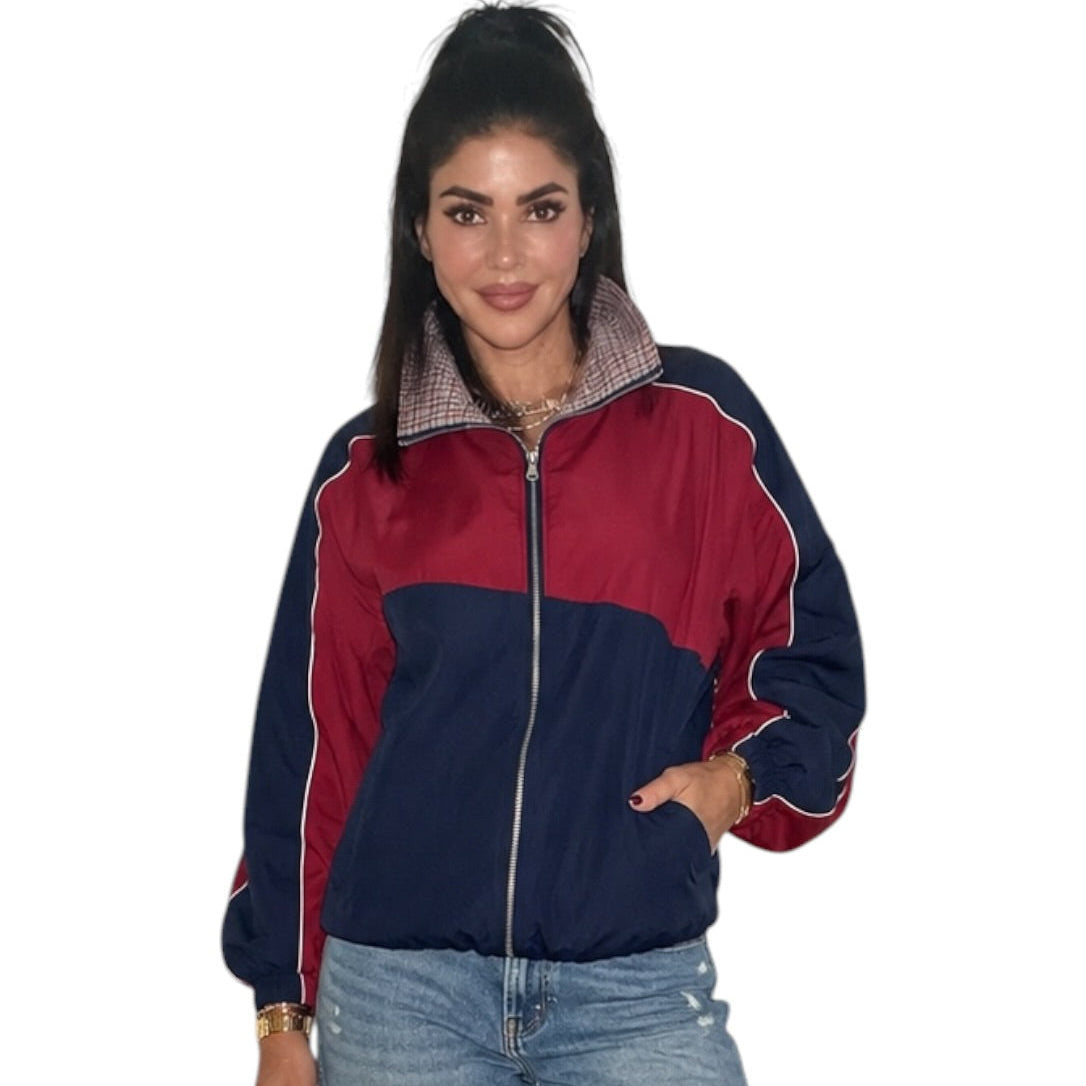 A woman modeling a colorblock windbreaker jacket with a navy blue lower body, burgundy red upper body and sleeves, and a plaid collar, paired with distressed light-wash blue jeans against a plain white background.