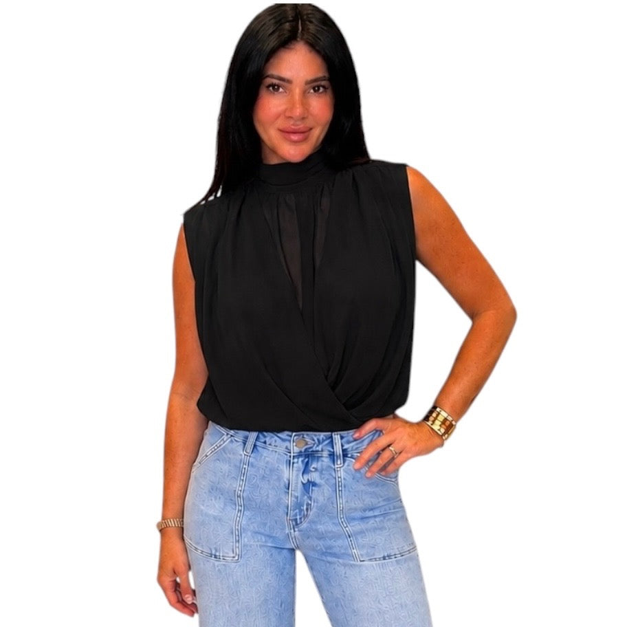 A woman modeling a sleeveless black blouse with a high mock neck and twisted front detail, paired with light blue jeans, against a plain white background.