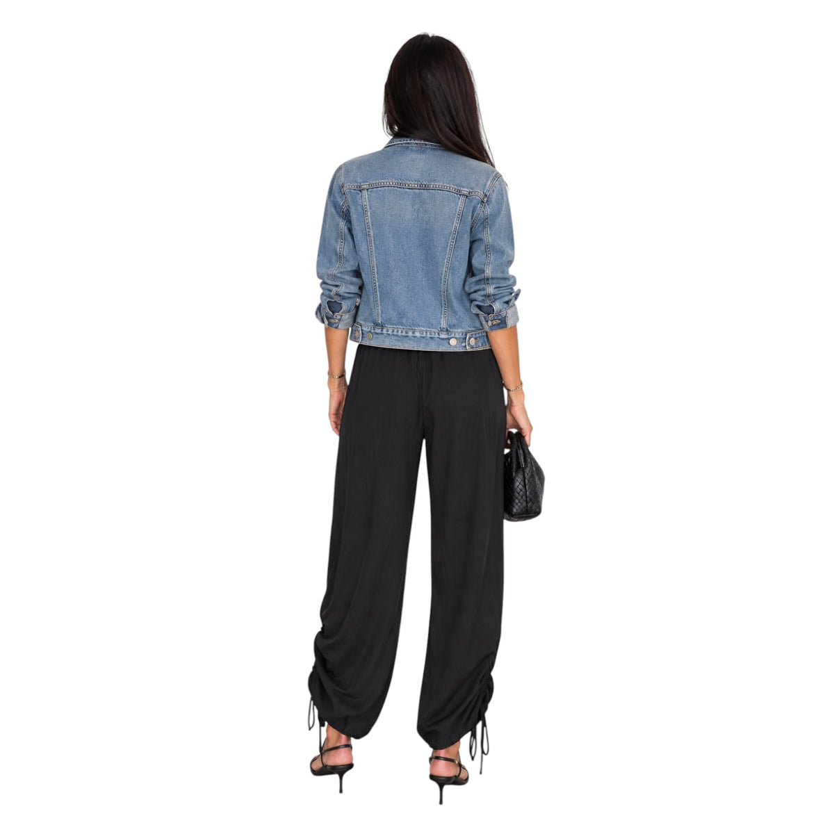 A woman shown from the back wearing a cropped light blue denim jacket, black wide-leg draped pants, and black heeled sandals, holding a small black handbag against a plain white background.