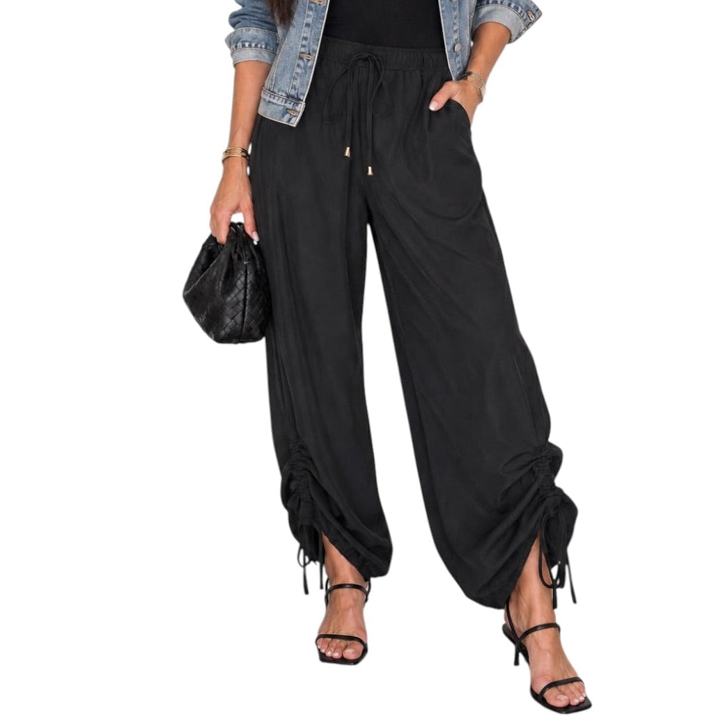 A woman modeling a black wide-leg pants with a drawstring waist and side tie details, styled with a light blue denim jacket, black tank top, and black strappy heels against a plain white background.