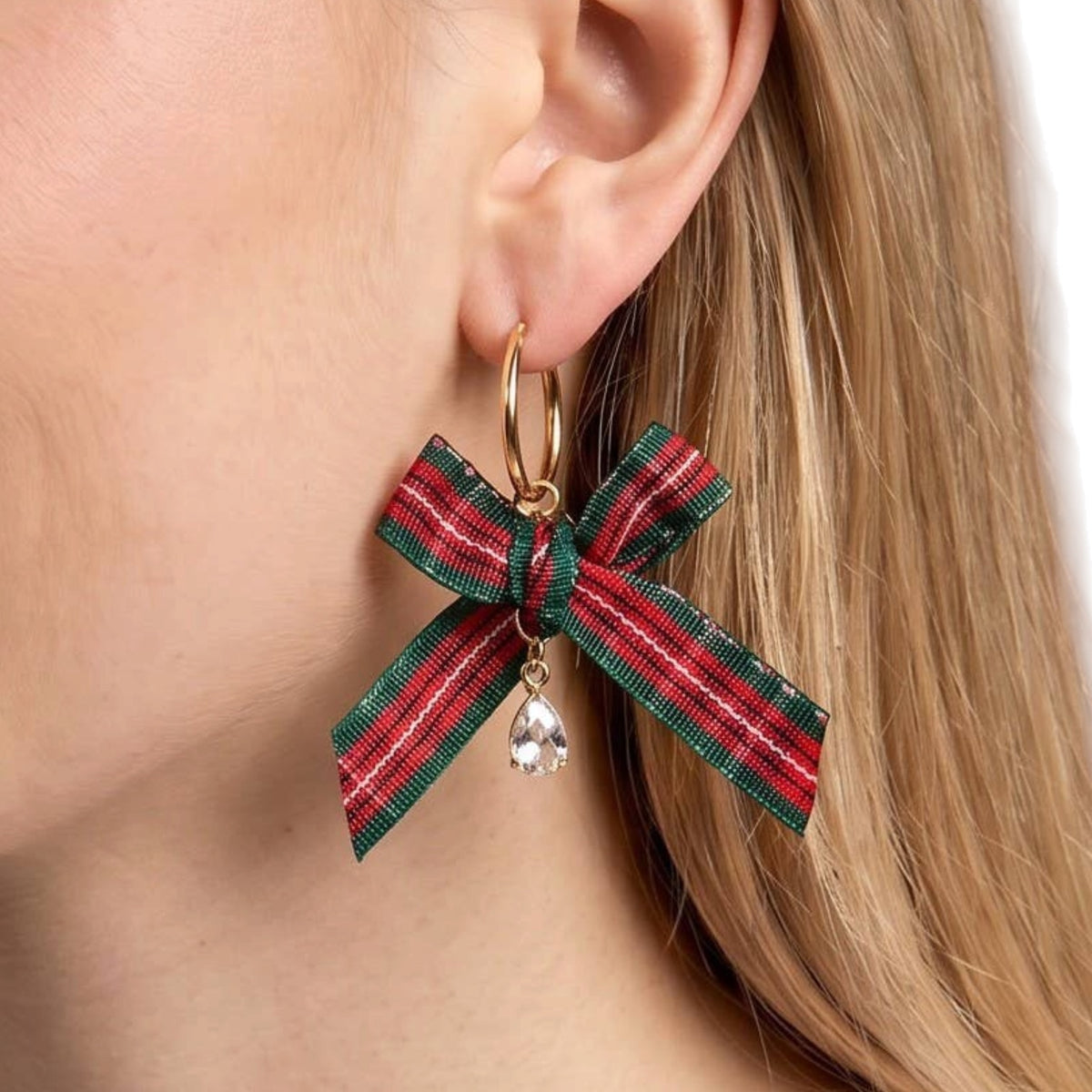 A close-up of a model's ear wearing a gold hoop earring with a red and green plaid ribbon bow charm and a clear teardrop crystal pendant.