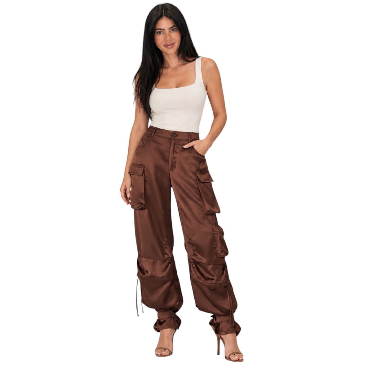 A woman modeling a pair of brown wide-leg cargo pants with multiple utility pockets, paired with a white tank top and nude high-heeled sandals, set against a plain white background.