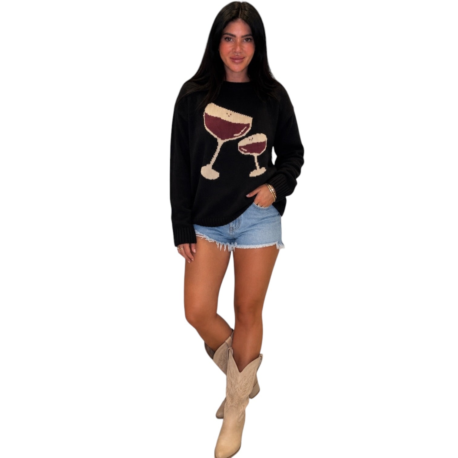 A woman modeling a black long-sleeve crewneck sweatshirt with a graphic of two wine glasses filled with red wine, styled with denim shorts and beige cowboy boots against a plain white background.