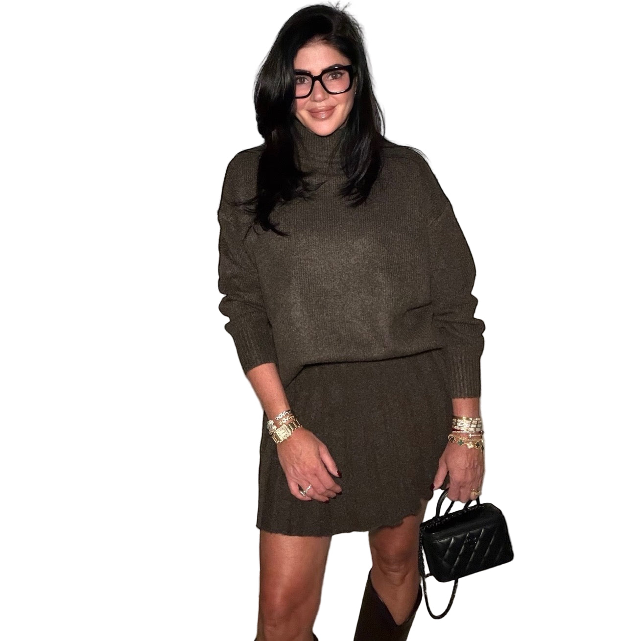 Woman wearing a brown sweater skirt set and holding a black handbag against a plain wall.