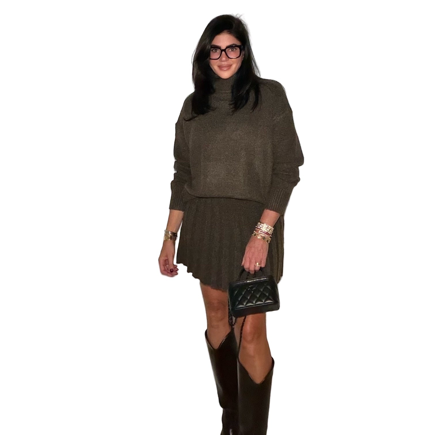 Woman wearing a dark green sweater and skirt set with black knee-high boots, holding a black handbag against a plain wall.