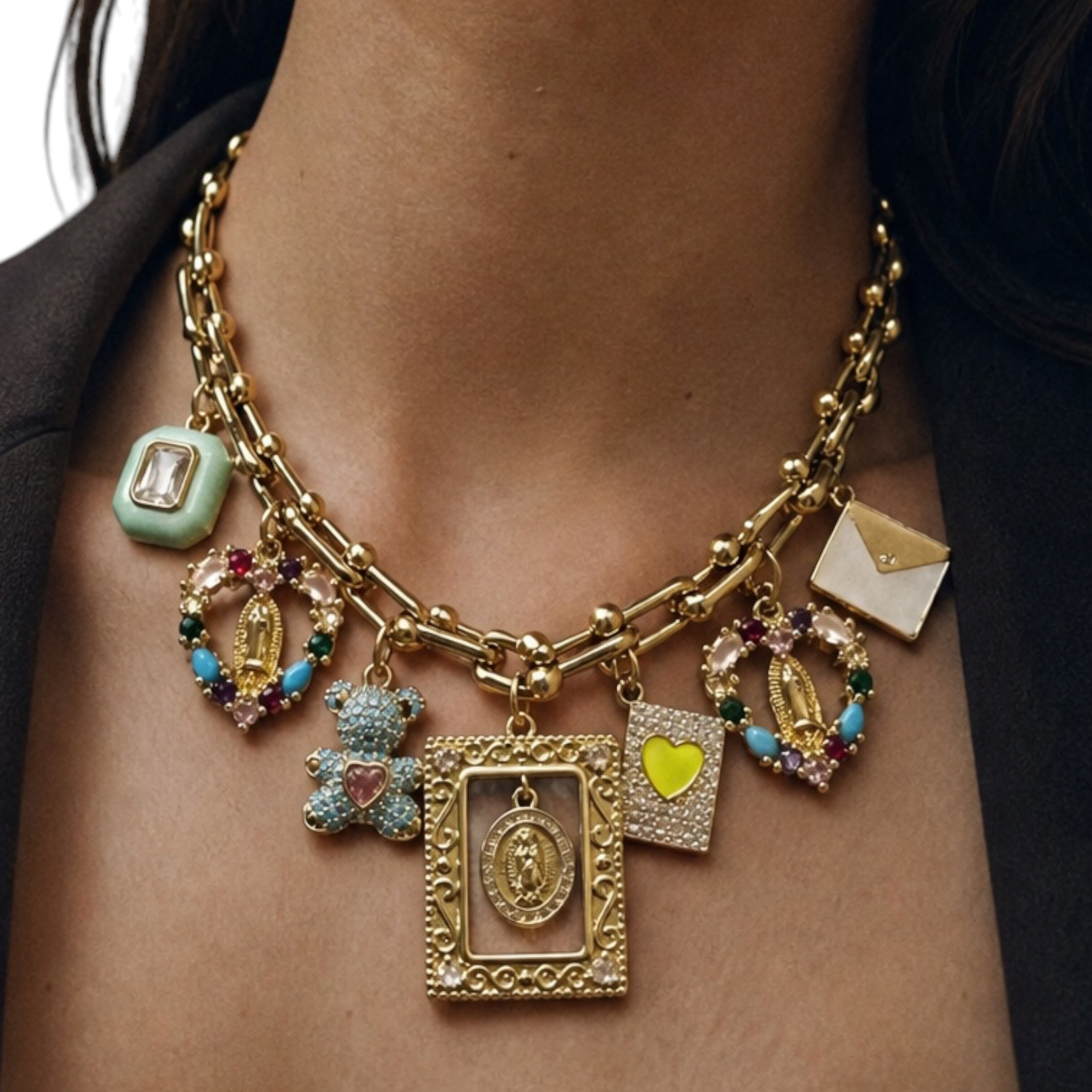 A close-up of a model wearing a gold charm necklace with multiple decorative pendants, including religious icons, a teddy bear, a heart, and an envelope charm.