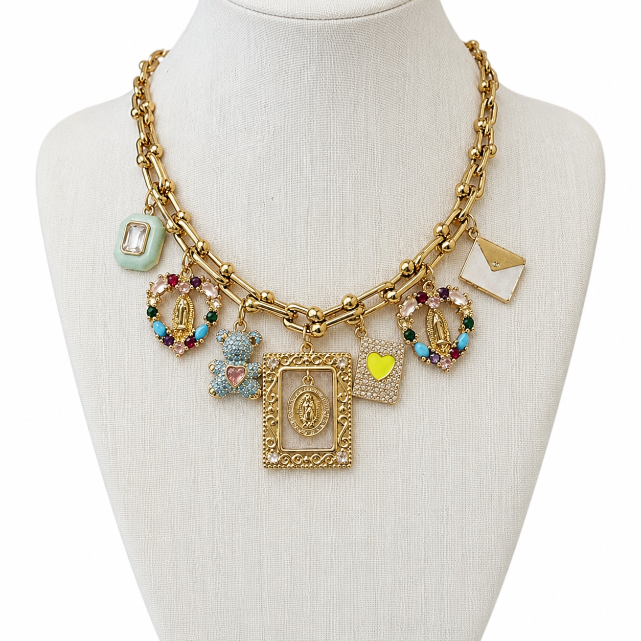 A gold-tone charm necklace displayed on a cream-colored mannequin bust. The necklace features a chunky link chain and multiple dangling charms, including two gold-tone Miraculous Medal pendants, a teddy bear charm, a small yellow heart, a light green square charm, and a white envelope charm.