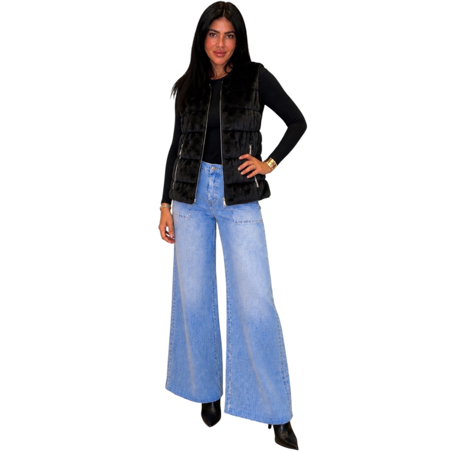 A woman modeling a black faux-fur vest over a black long-sleeve top and light blue wide-leg denim jeans, set against a plain white background.