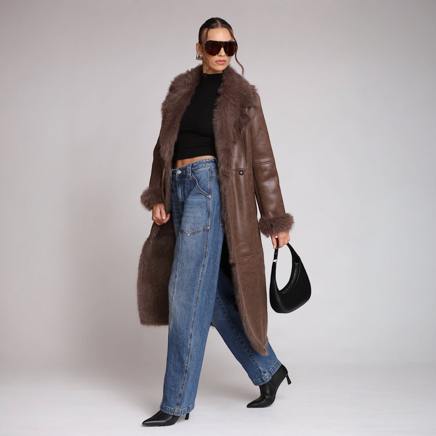 Woman wearing a long brown fur-trimmed coat, black top, blue jeans, and black shoes on a plain background.