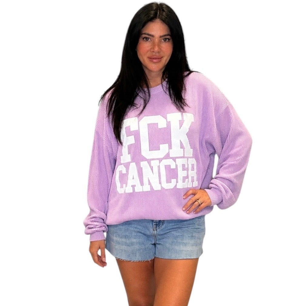A woman modeling a lavender purple ribbed knit sweatshirt with 'FCK CANCER' printed in bold white block letters across the front, paired with denim shorts against a plain white background.