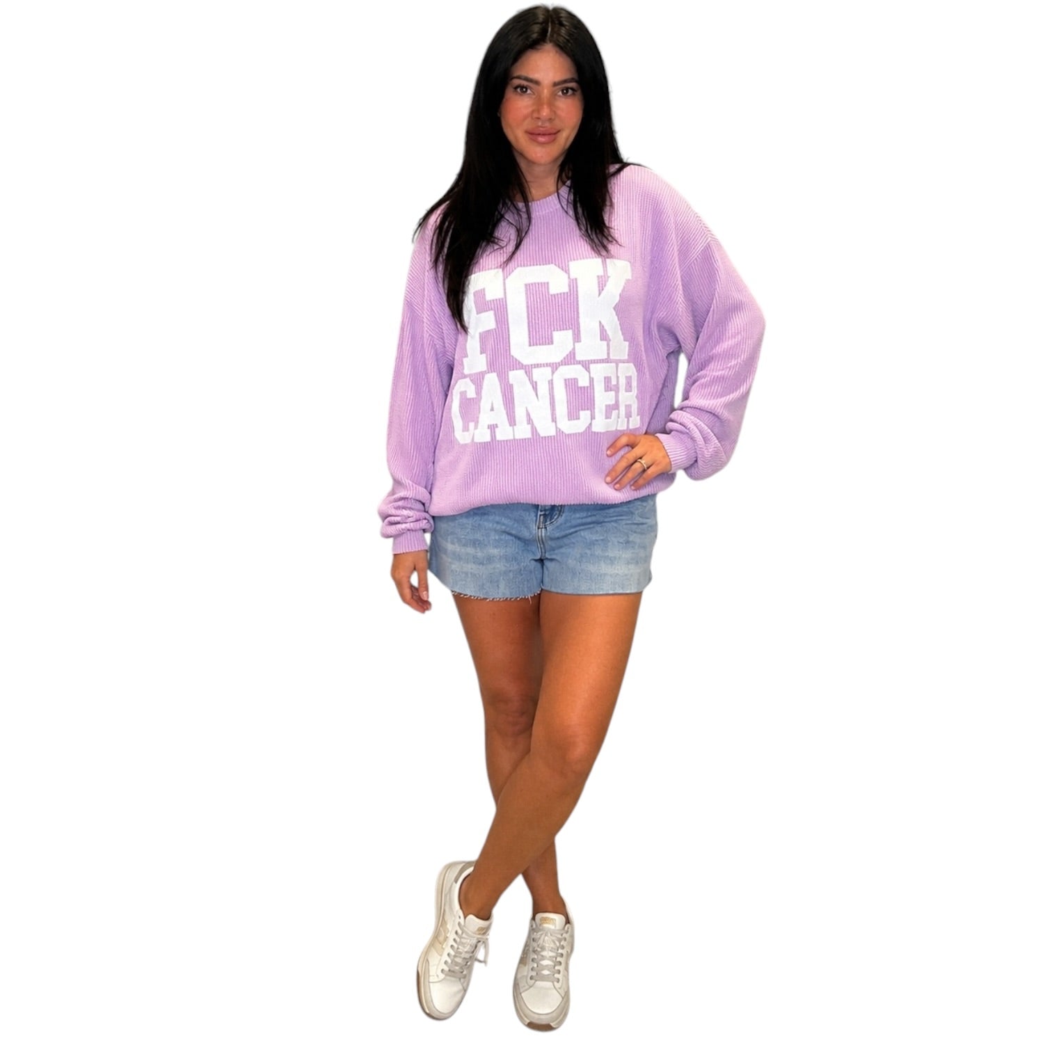 A woman modeling a lavender purple sweatshirt with 'FCK CANCER' printed in white block letters, paired with denim shorts and white sneakers, against a plain white background.