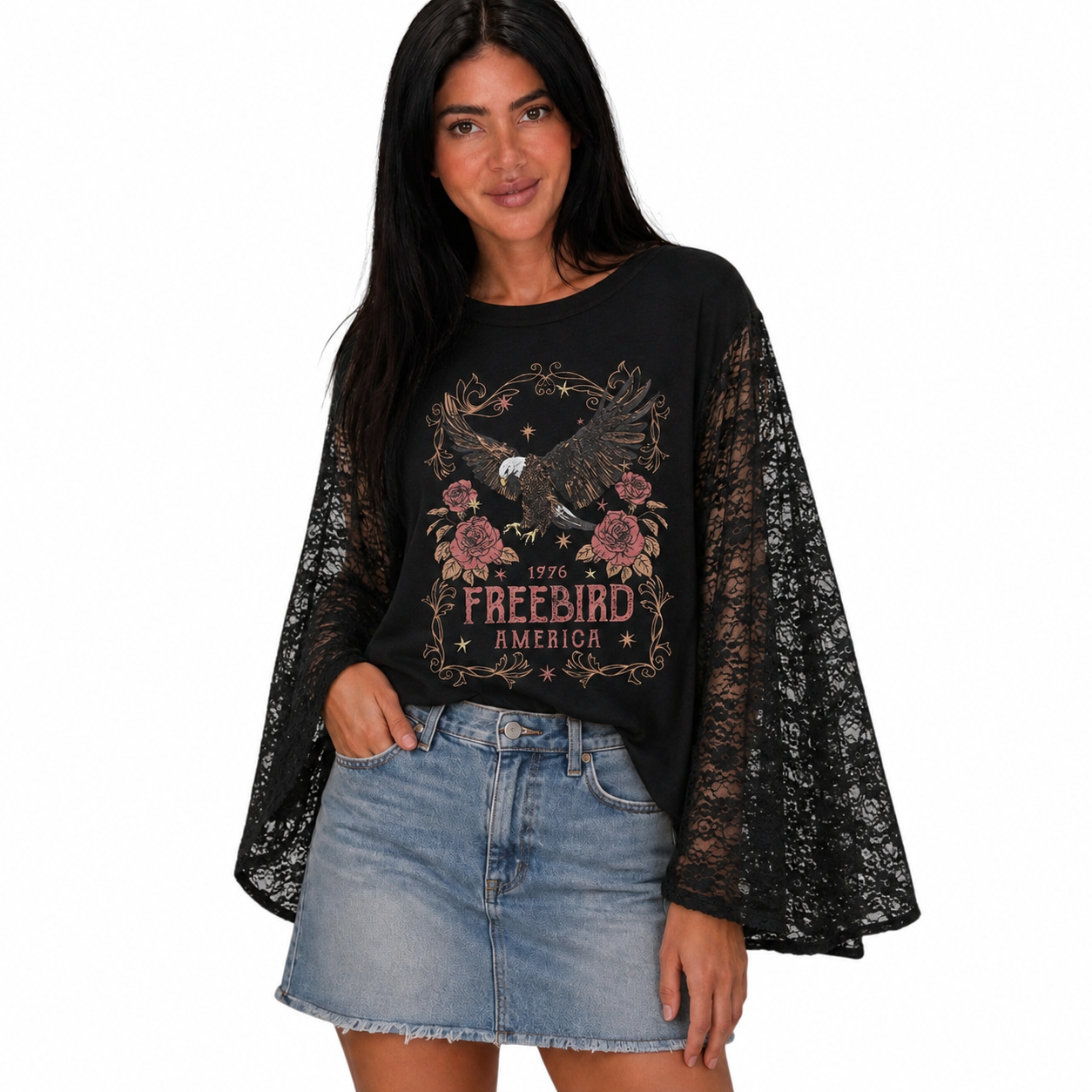 A woman modeling a black long-sleeve t-shirt with sheer black lace bell sleeves and a graphic print featuring an eagle, flowers, and the text '1976 FREEBIRD AMERICA'. She is wearing a light-wash denim mini skirt against a plain white background.