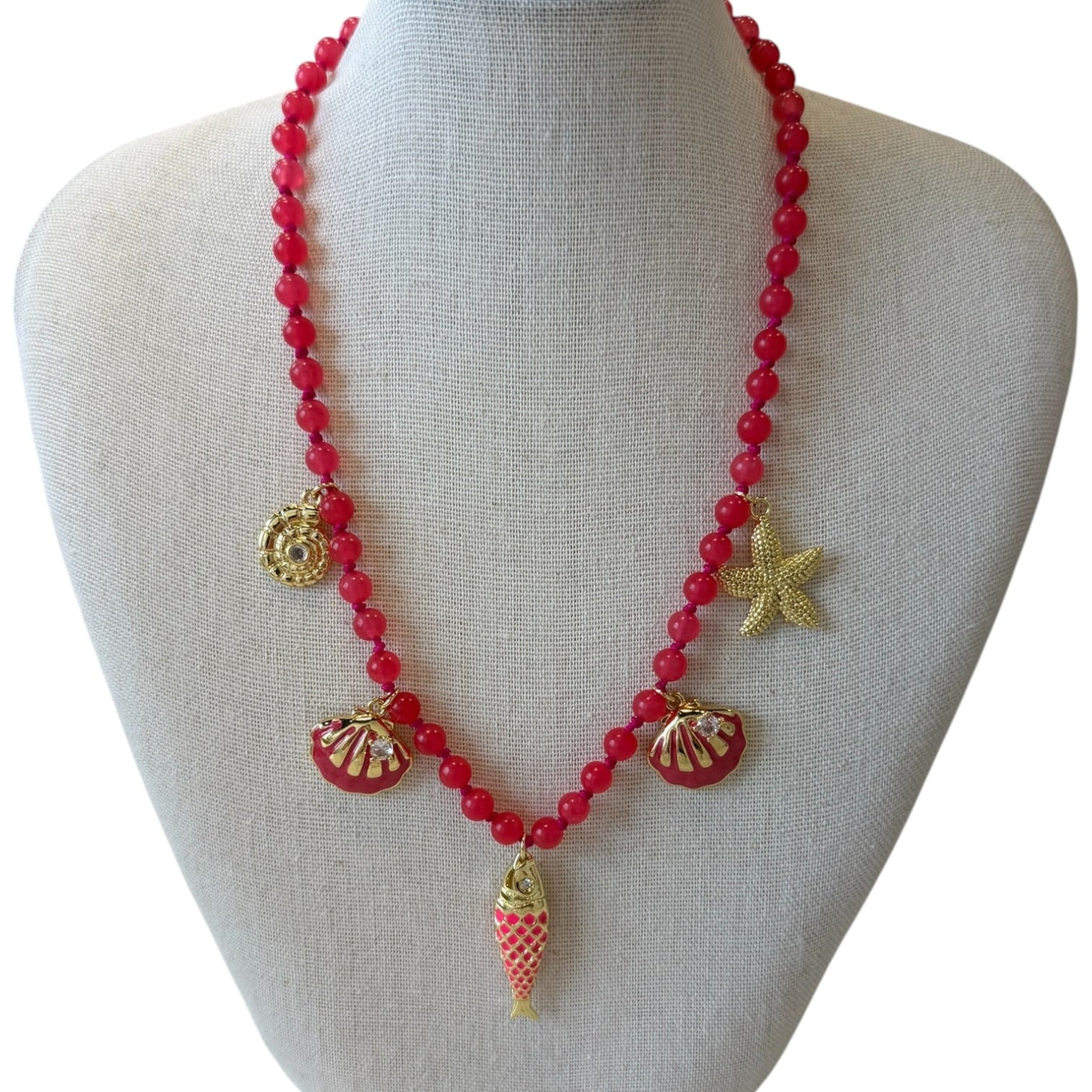 A red beaded necklace with gold-toned seashell, starfish, and fish charms, displayed on a light gray fabric mannequin bust.