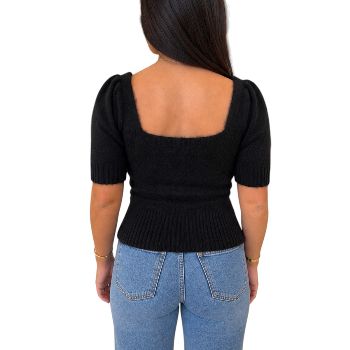 A woman viewed from the back wearing a black short-sleeve knit top with a square neckline and puff sleeves, paired with light blue jeans, against a plain white background.