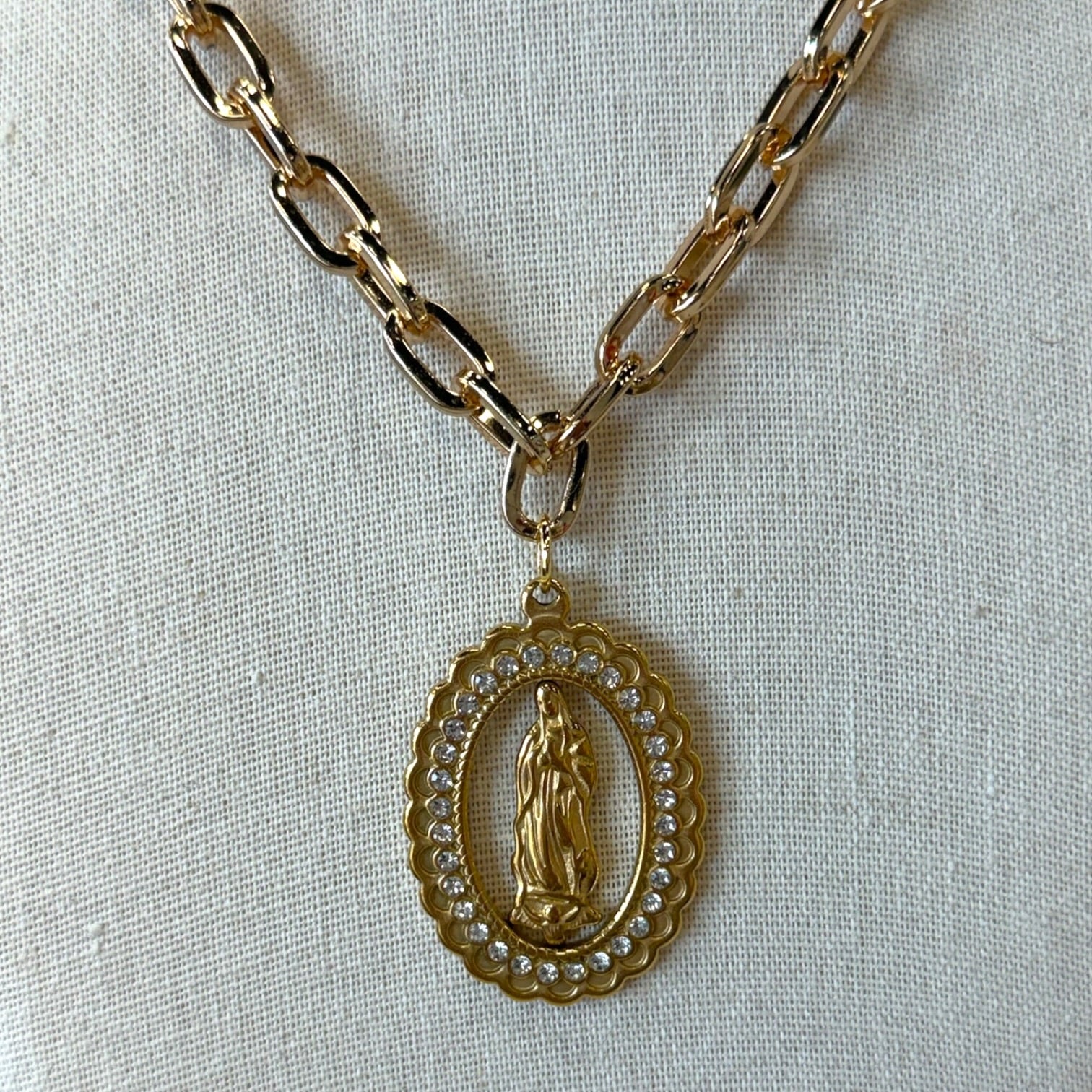 Gold chain necklace with an oval pendant featuring a religious figure surrounded by small clear stones, displayed on a textured off-white fabric background.