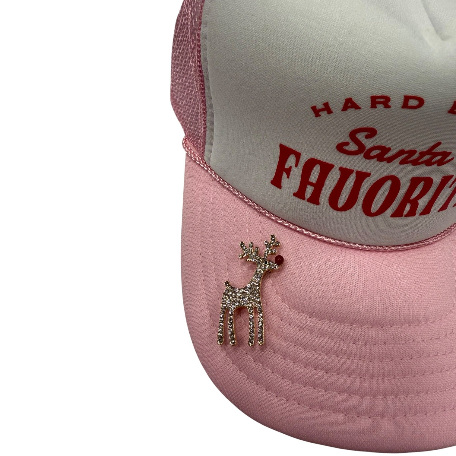 A close-up of a pink and white trucker hat featuring red embroidered text on the front panel and a rhinestone-embellished reindeer pin on the brim.