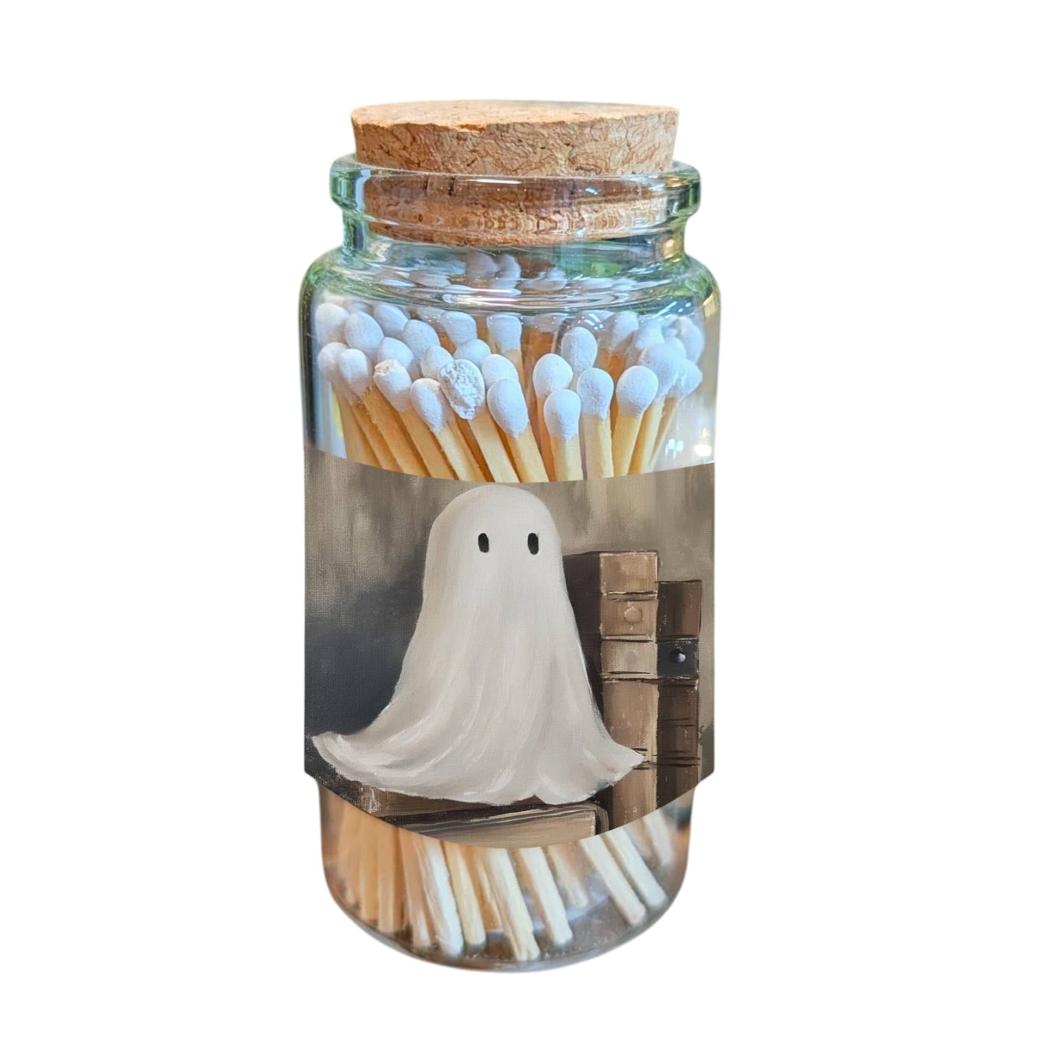 A clear glass jar with a cork stopper, filled with matchsticks and featuring a printed label of a white ghost standing in front of old books.