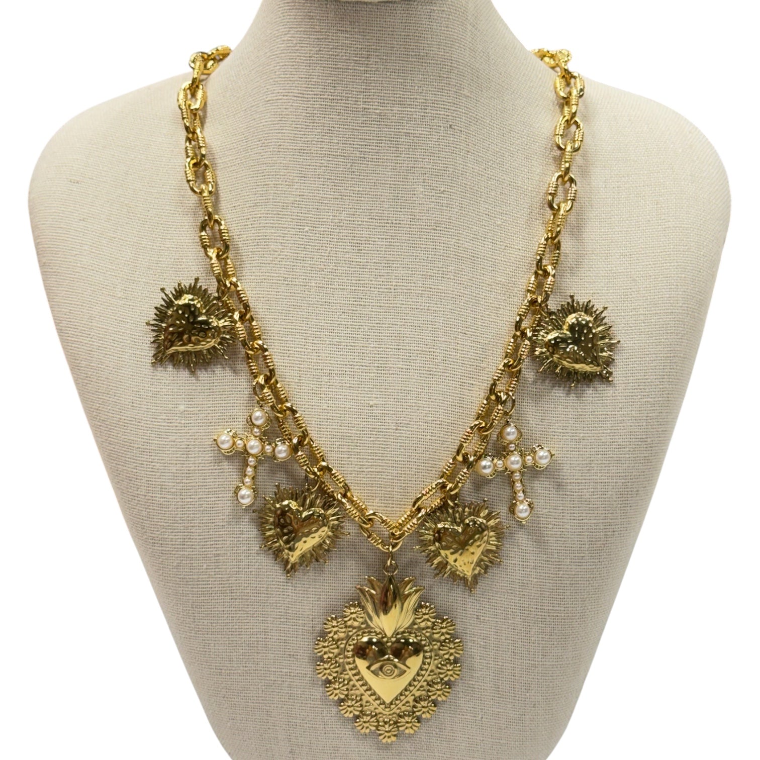 A gold-toned statement necklace displayed on a beige fabric mannequin bust. The necklace features a large Sacred Heart pendant, smaller heart charms, textured sunburst details, and small pearl-like bead accents.