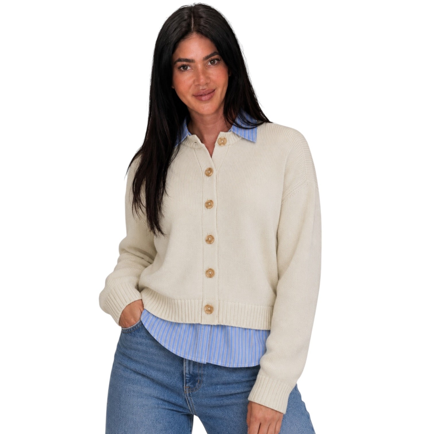 A woman modeling a cream-colored knit button-up cardigan sweater with brown buttons, layered over a light blue and white striped shirt and paired with blue jeans, against a solid white background.