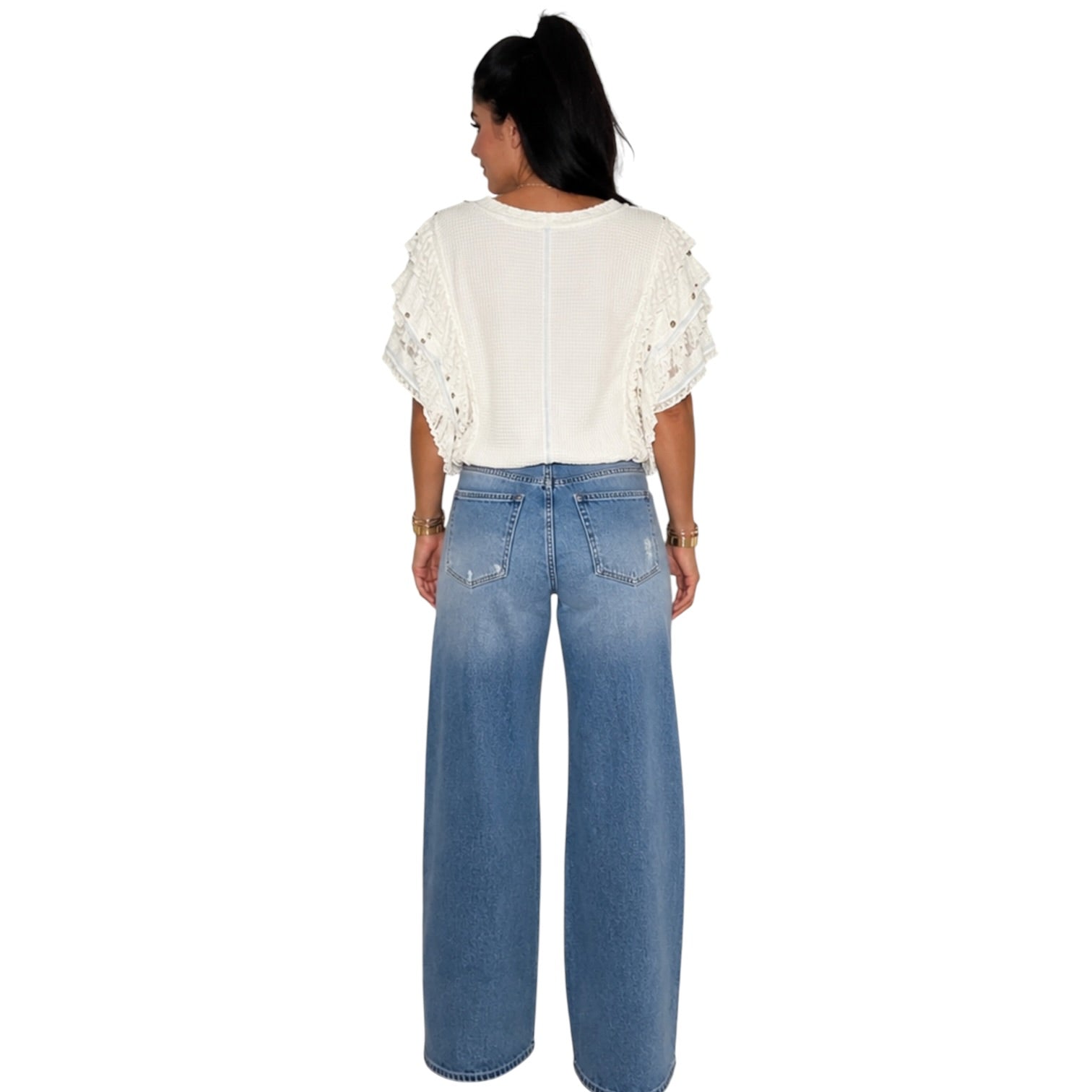 A woman shown from the back wearing a white short-sleeve top with textured, eyelet-style ruffle sleeves and high-waisted, wide-leg blue jeans, set against a plain white background.