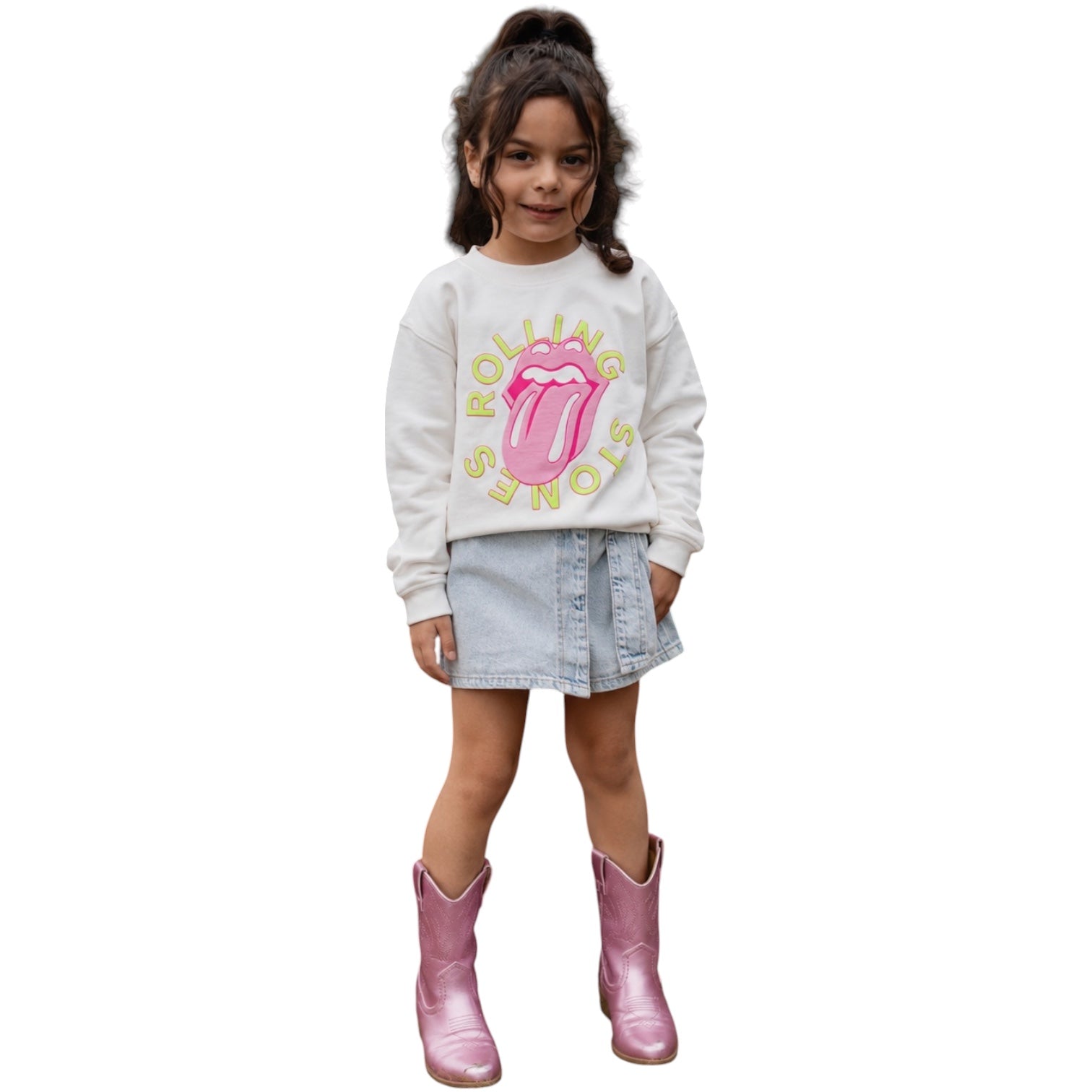 A young girl stands against a plain white background wearing a white long-sleeve sweatshirt with a pink Rolling Stones tongue graphic and yellow text, a light blue denim skirt, and shiny pink cowboy boots.
